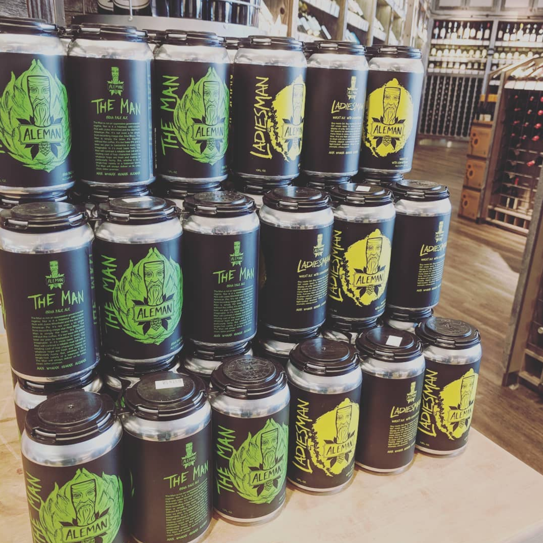 Stacked cans of Aleman beer, including The Man and Ladiesman varieties, on a table in a store.