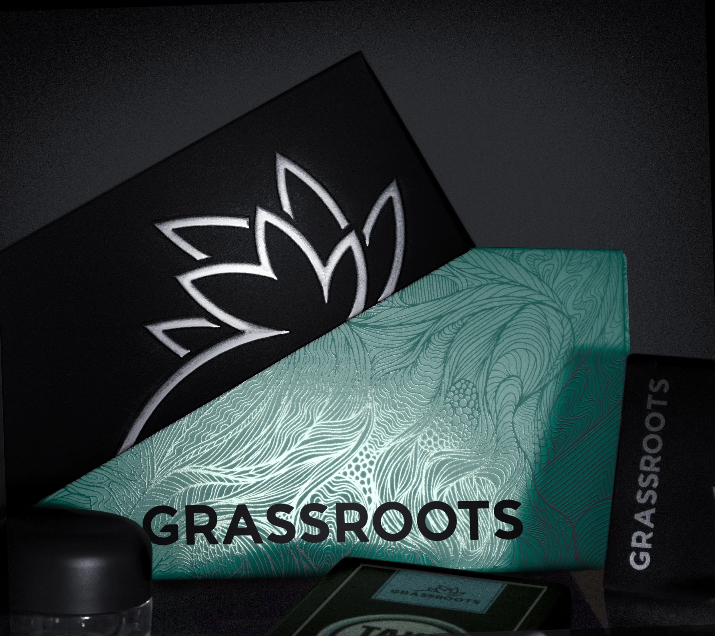 A black box with a white stylized lotus flower logo, a teal box with intricate line patterns and the word "GRASSROOTS" in black, a small jar with a black lid, and a partially visible green and black item.