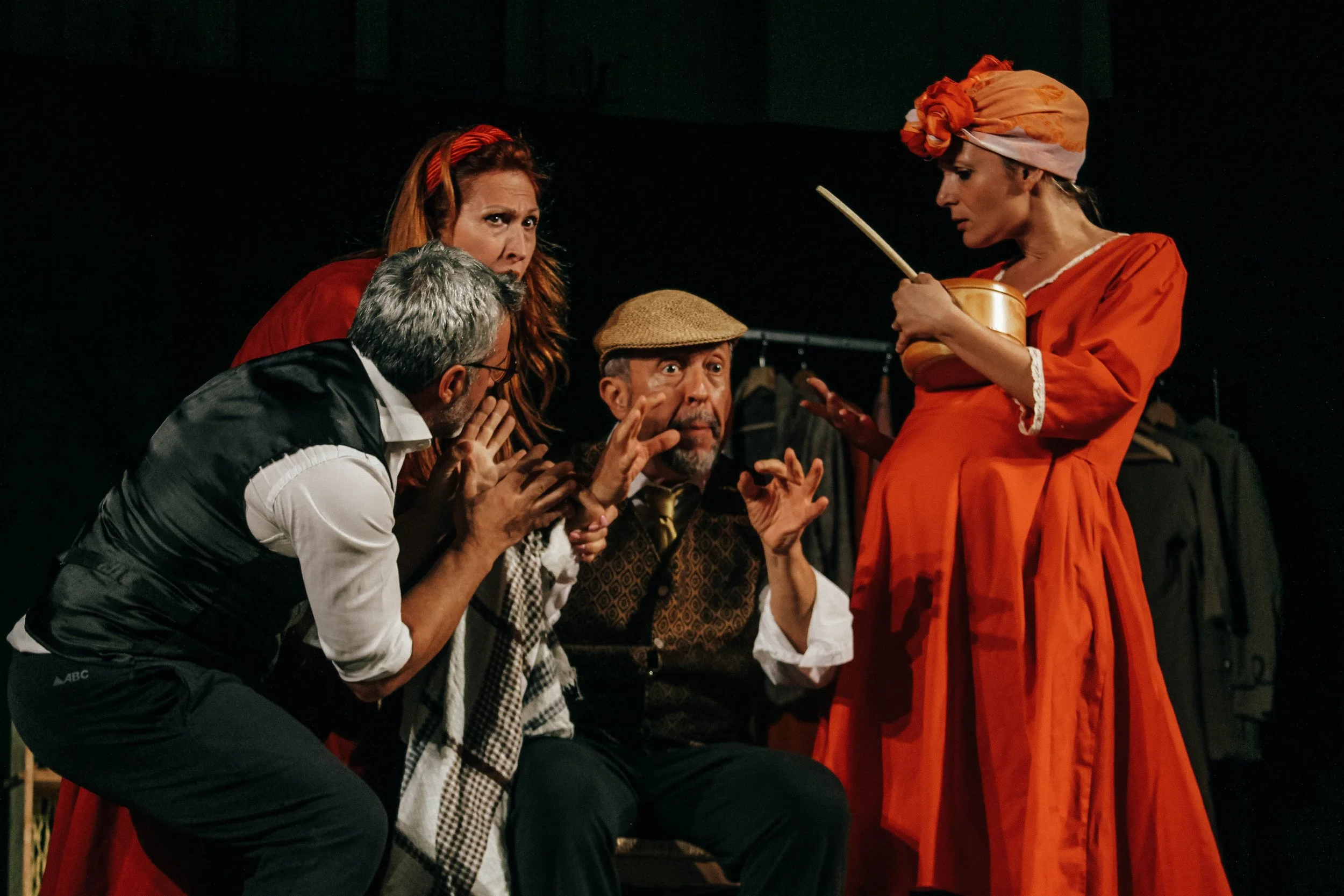 Four actors performing on stage in costume, with one woman pregnant, and three others engaged in a theatrical scene. The woman on the right is dressed in an orange dress and headscarf, holding a pot and a spoon. The group appears to be intense, with exaggerated expressions, suggesting a dramatic or comedic play.