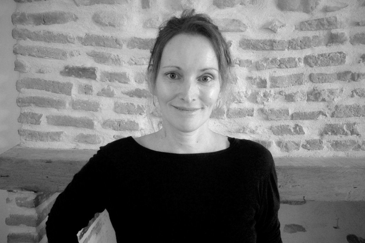 A woman with light skin and brown hair tied back, smiling, wearing a black top, standing in front of a brick wall with a wooden surface behind her.