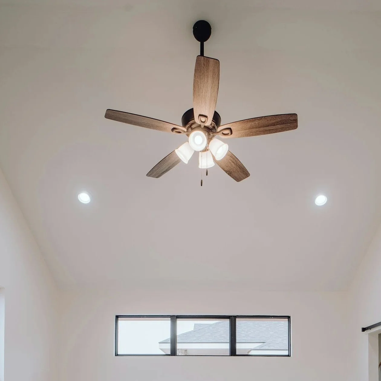 Ceiling fan with wooden blades and integrated lighting in a modern room with white walls, recessed lighting, and a horizontal window.