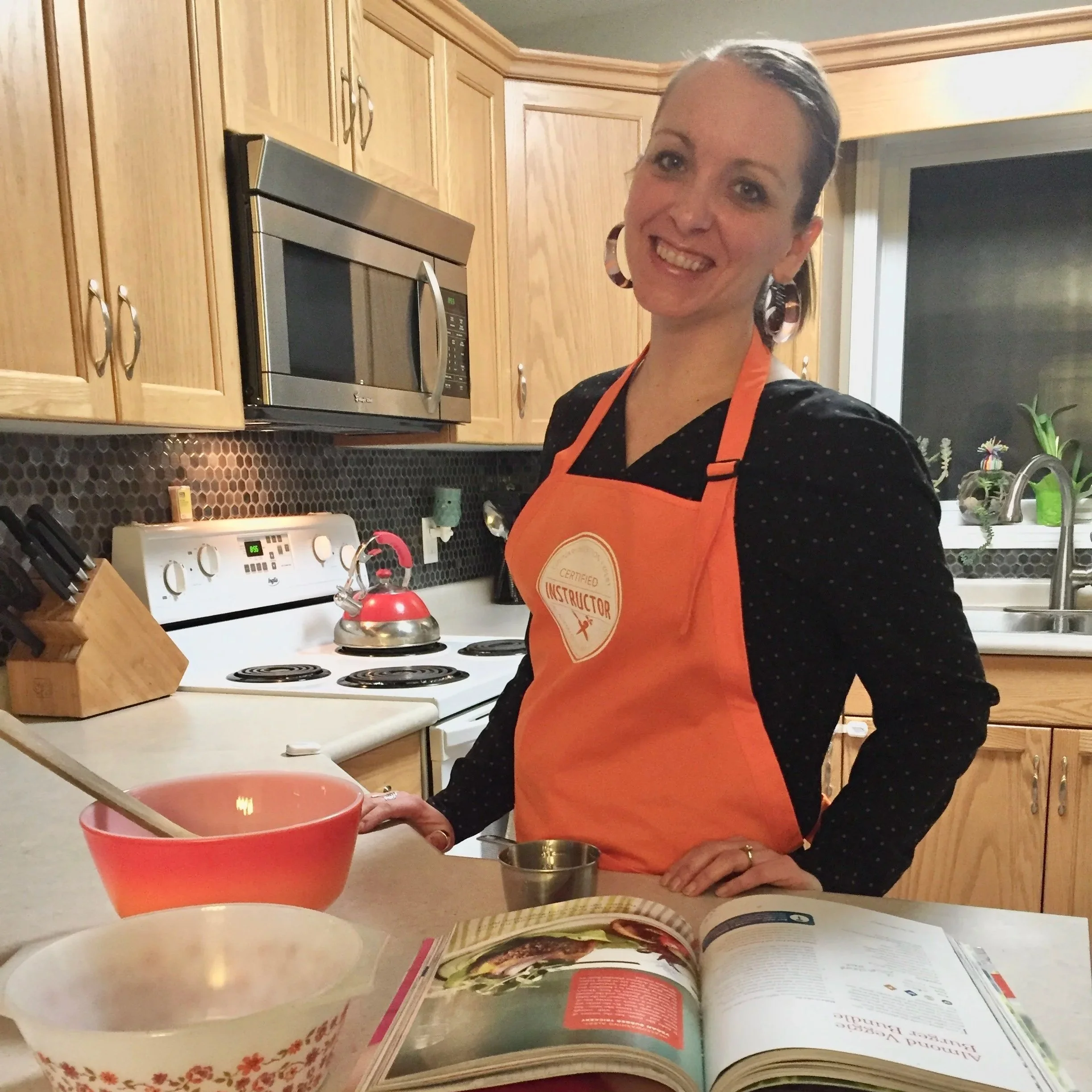 Megan Deaust wearing an orange apron with a 'Culinary Nutrition Expert Certified Instructor' badge smiles in a kitchen. She stands at a counter with a cookbook open in front of her, a pink mixing bowl, and a measuring cup arranged nearby.