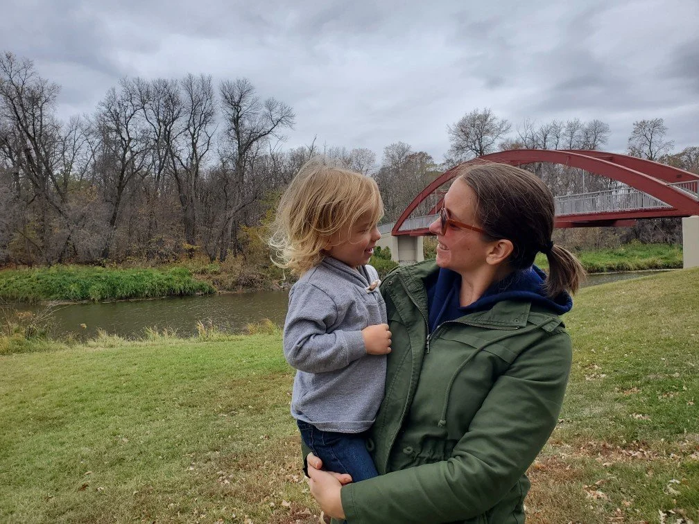 Megan Deaust with sunglasses holding her son in an outdoor park near a river and a red-covered bridge on a cloudy day.