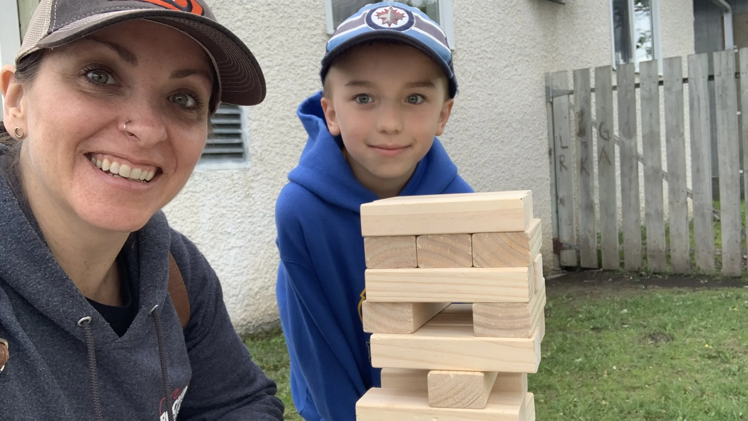 Megan Deaust and her son smiling and playing Jenga outdoors on grass, with a wall and wooden fence in the background.