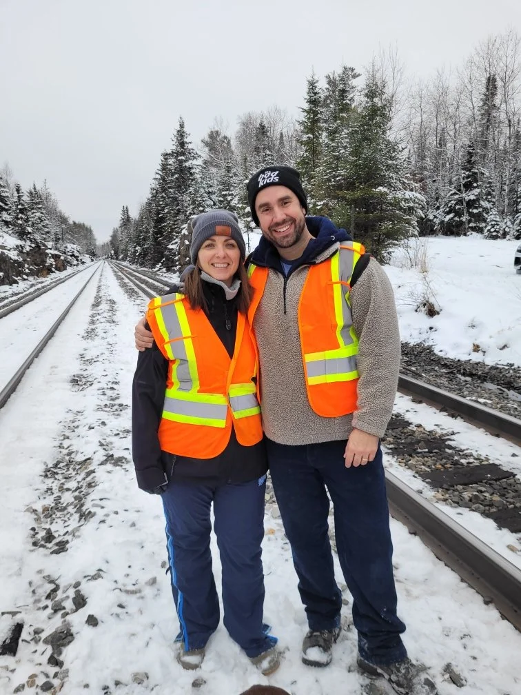 Megan Deaust and Trevor Martens wearing safety vests stand on snow-covered railroad tracks in a winter forest.