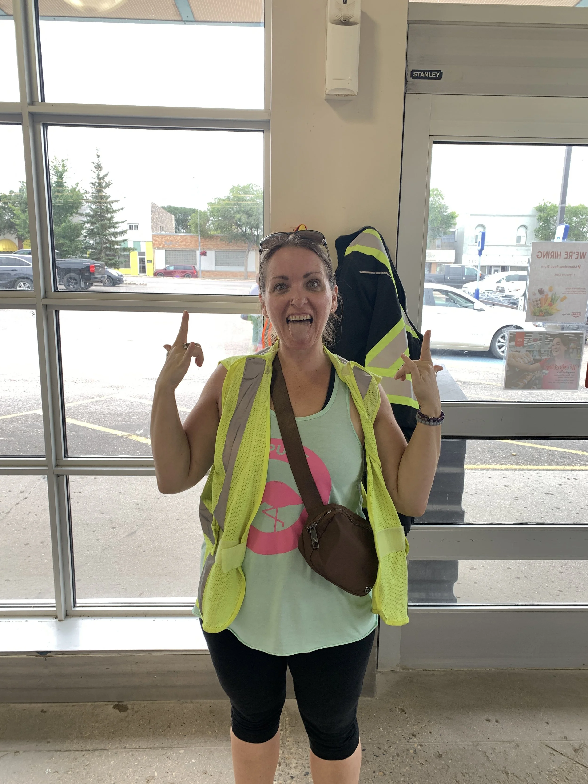 Megan Deaust standing indoors in front of large windows, wearing a neon yellow safety vest, black leggings, a tank top, and a brown crossbody bag. She is smiling with her tongue out and making rock on hand gestures. Minnedosa, MB.