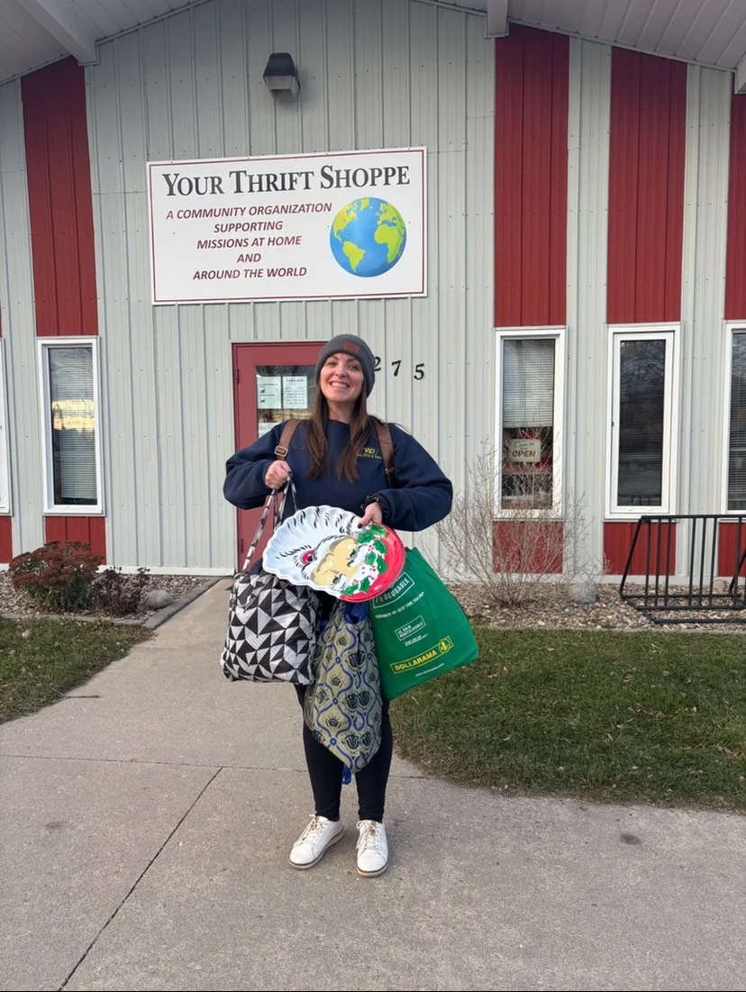 Megan Deaust wearing a gray toque, navy blue sweatshirt, and patterned pants, is standing outside a thrift store holding a Christmas-themed plate and shopping bags. The store has a sign reading 'Your Thrift Shoppe' and a globe graphic.