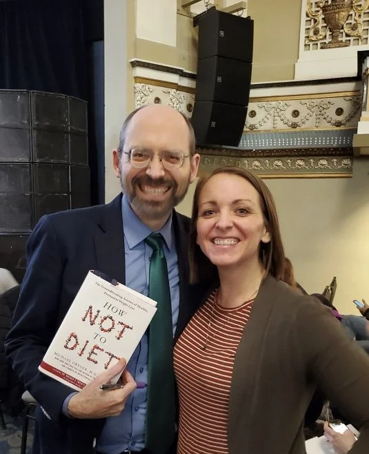 Megan Deaust and Dr. Michael Greger smiling and posing for a photo indoors; the man is holding a book titled 'How Not to Die'.
