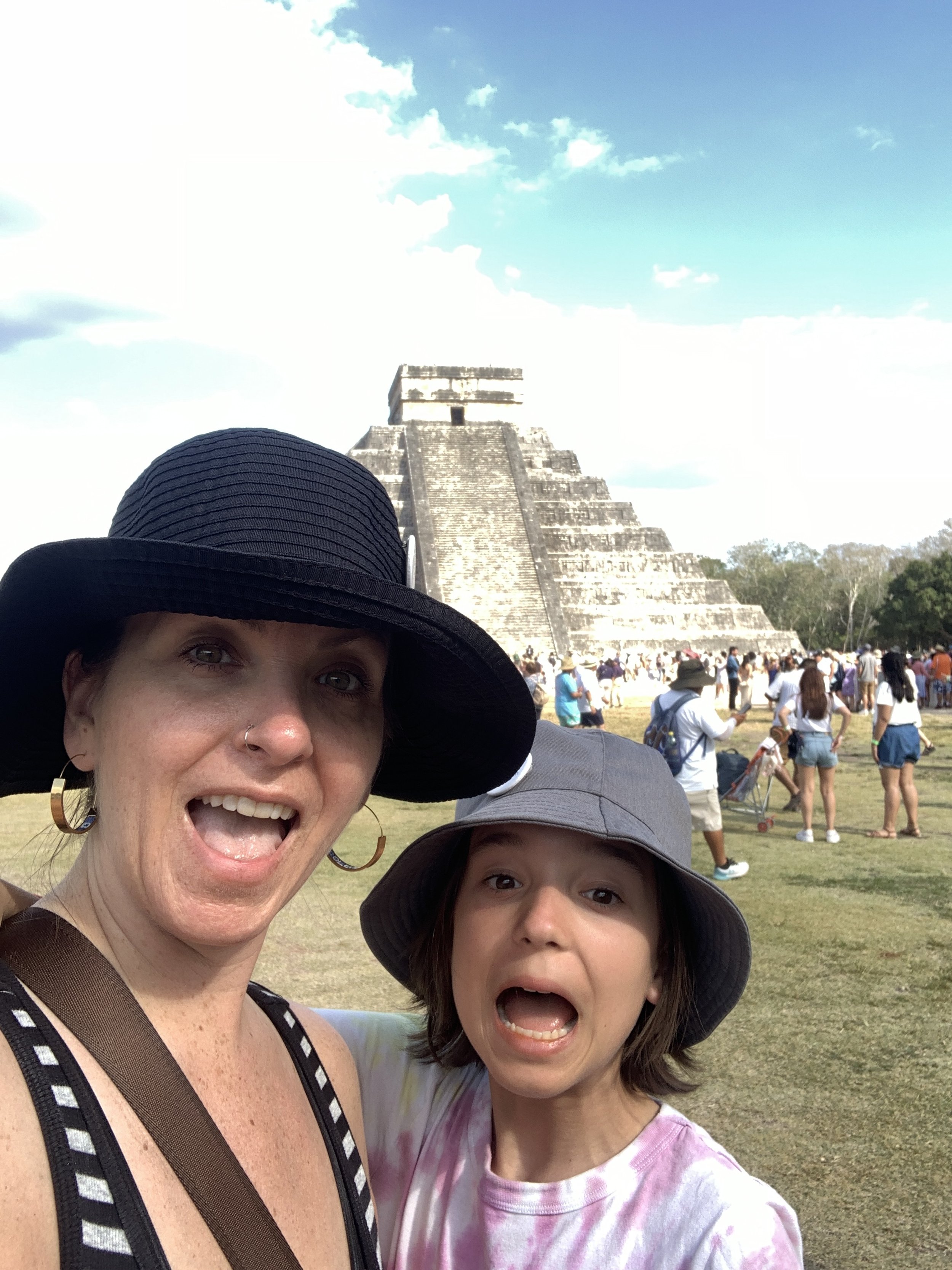 Megan Deaust and daughter taking a selfie in front of the ancient Mayan pyramid El Castillo at Chichen Itza, with many tourists in the background.
