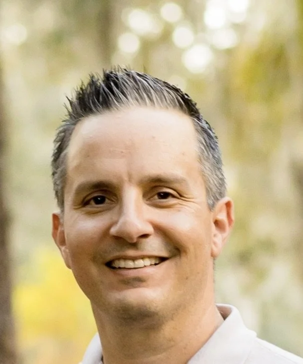 Close-up of a smiling man with short, dark hair in an outdoor setting with blurred green trees and sunlight in the background.