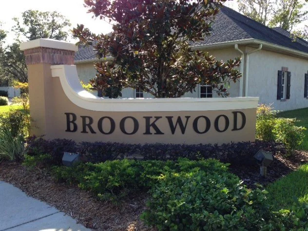 Sign reading 'Brookwood' surrounded by greenery and a house in the background.