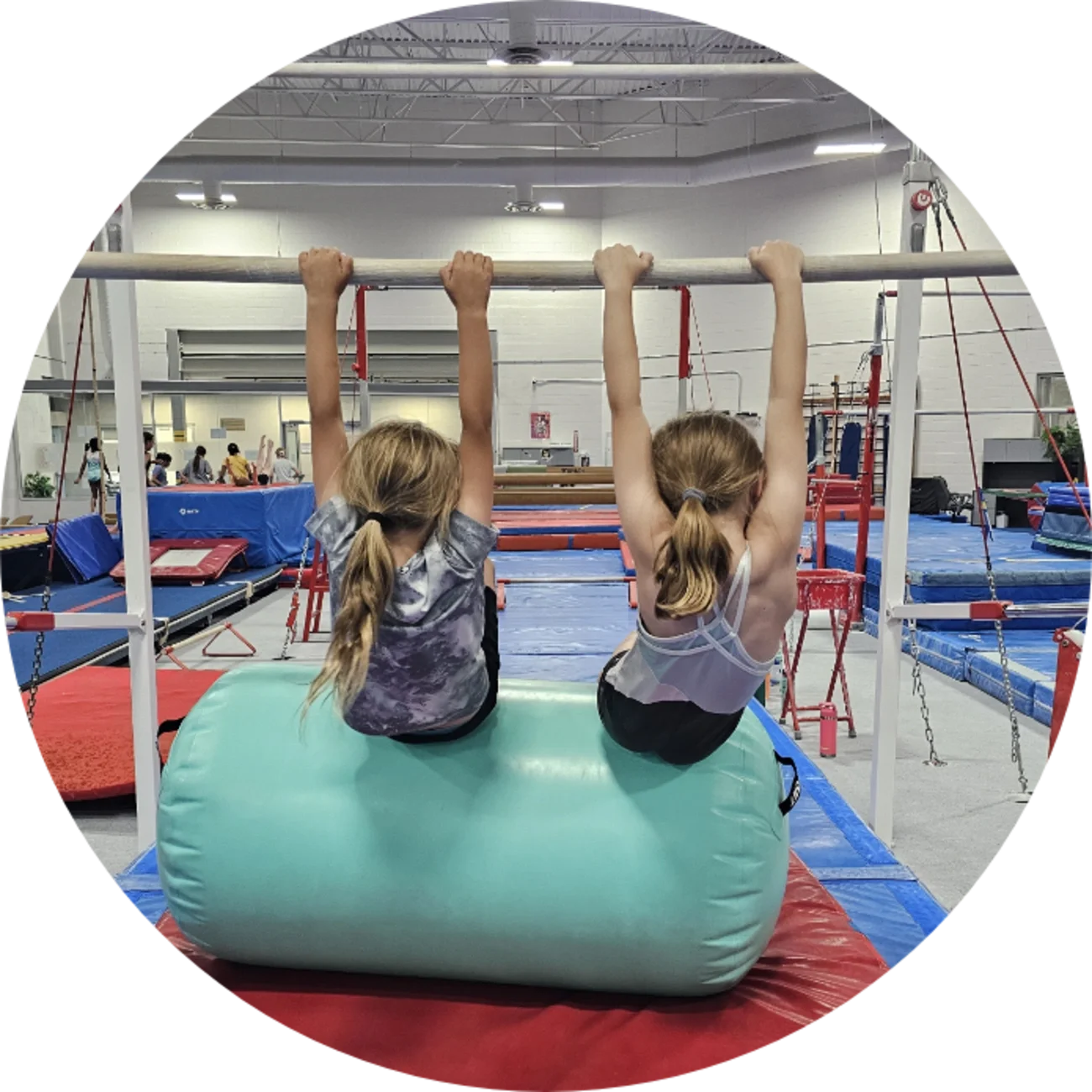Two young girls sitting on a large green foam block in a gymnastics gym, holding onto a horizontal bar as part of their gymnastics training.