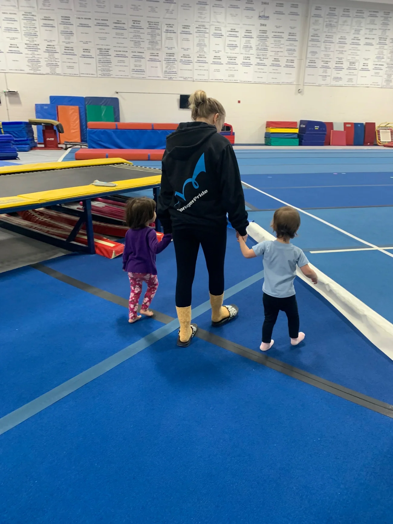 A woman holding hands with two young children walking inside a gymnastics gym with blue mats and gymnastics equipment.