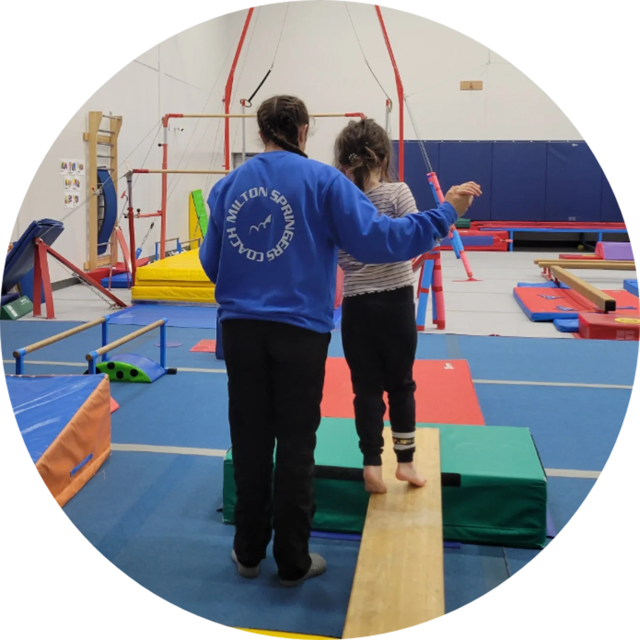 A girl balancing on a wooden beam with assistance from an instructor in a gymnastics training facility filled with equipment.