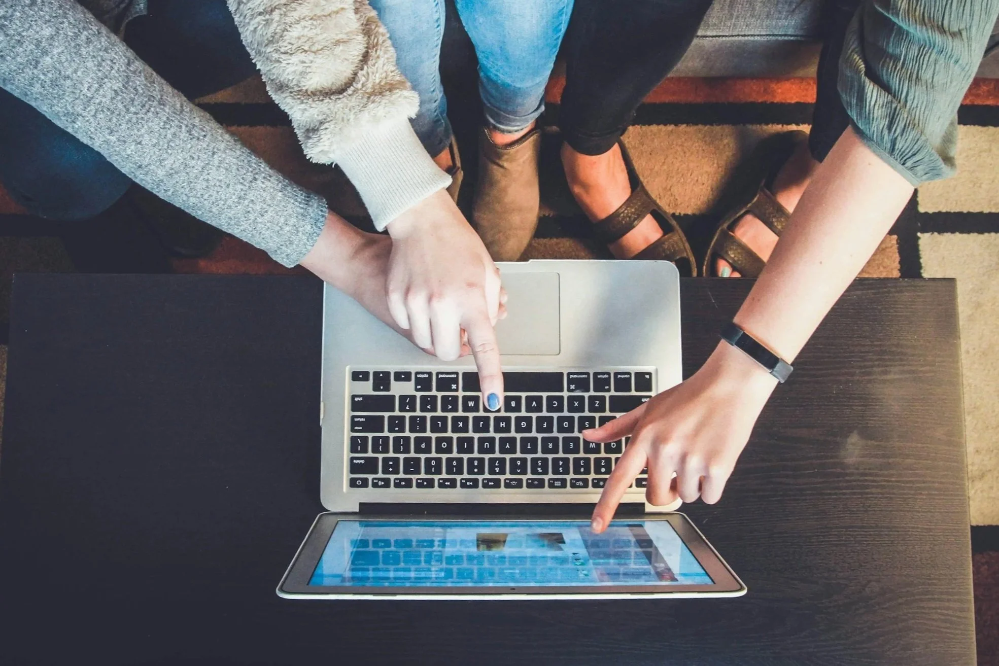 Three people sitting on a black table, using a MacBook and a tablet, with their fingers pointing at the screen.