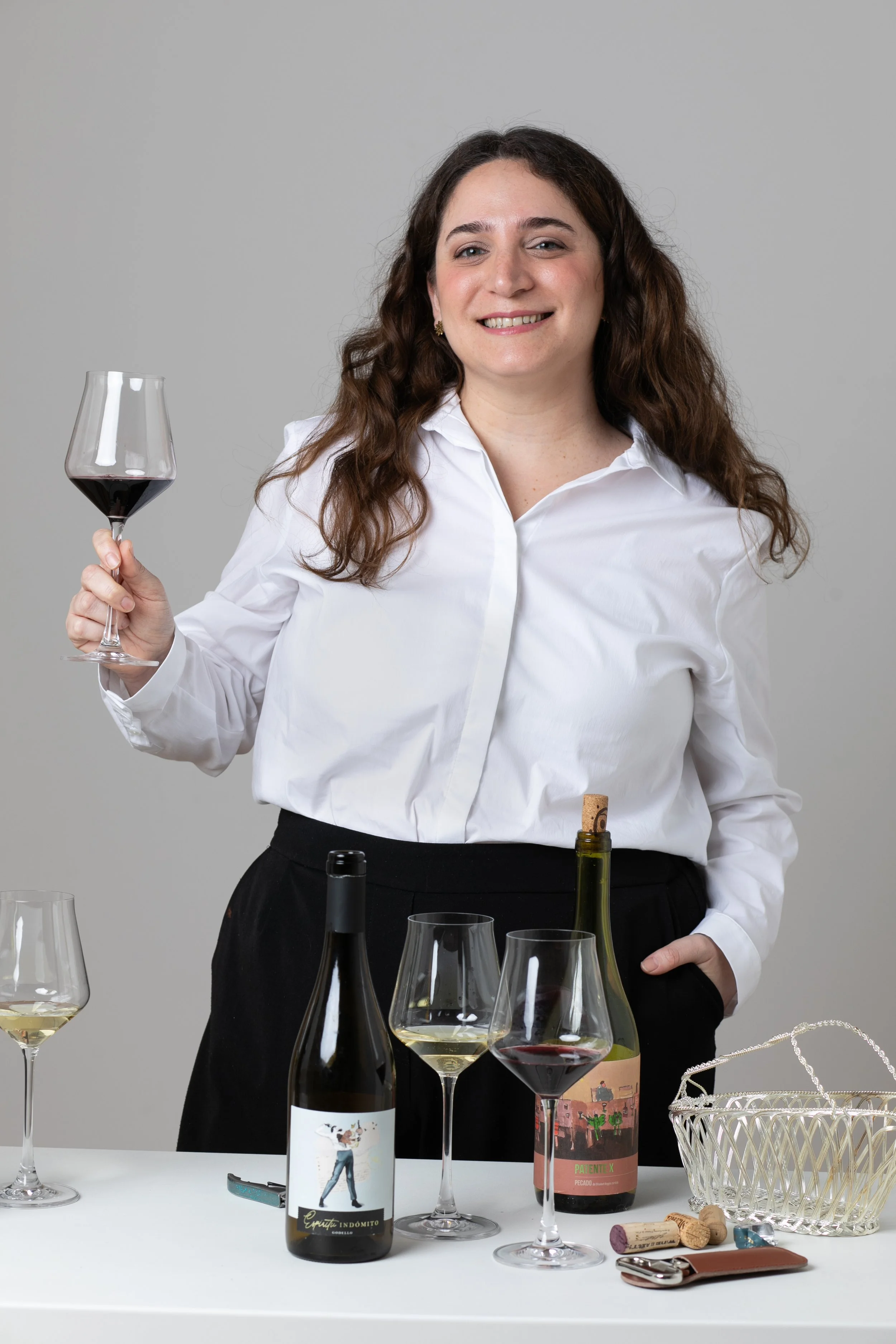 Mujer sonriendo y sosteniendo una copa de vino tinto, rodeada de varias copas y botellas de vino en una mesa.