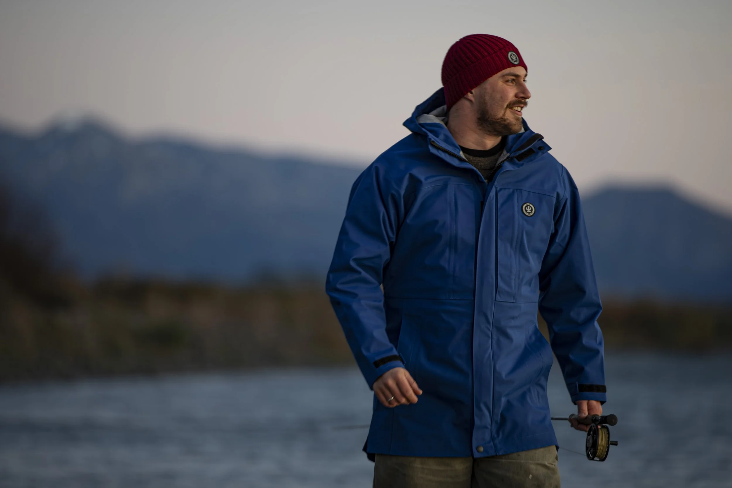 A man in a blue waterproof jacket and red knit hat holding a fishing rod outdoors, with mountains and a body of water in the background, during dusk or dawn.