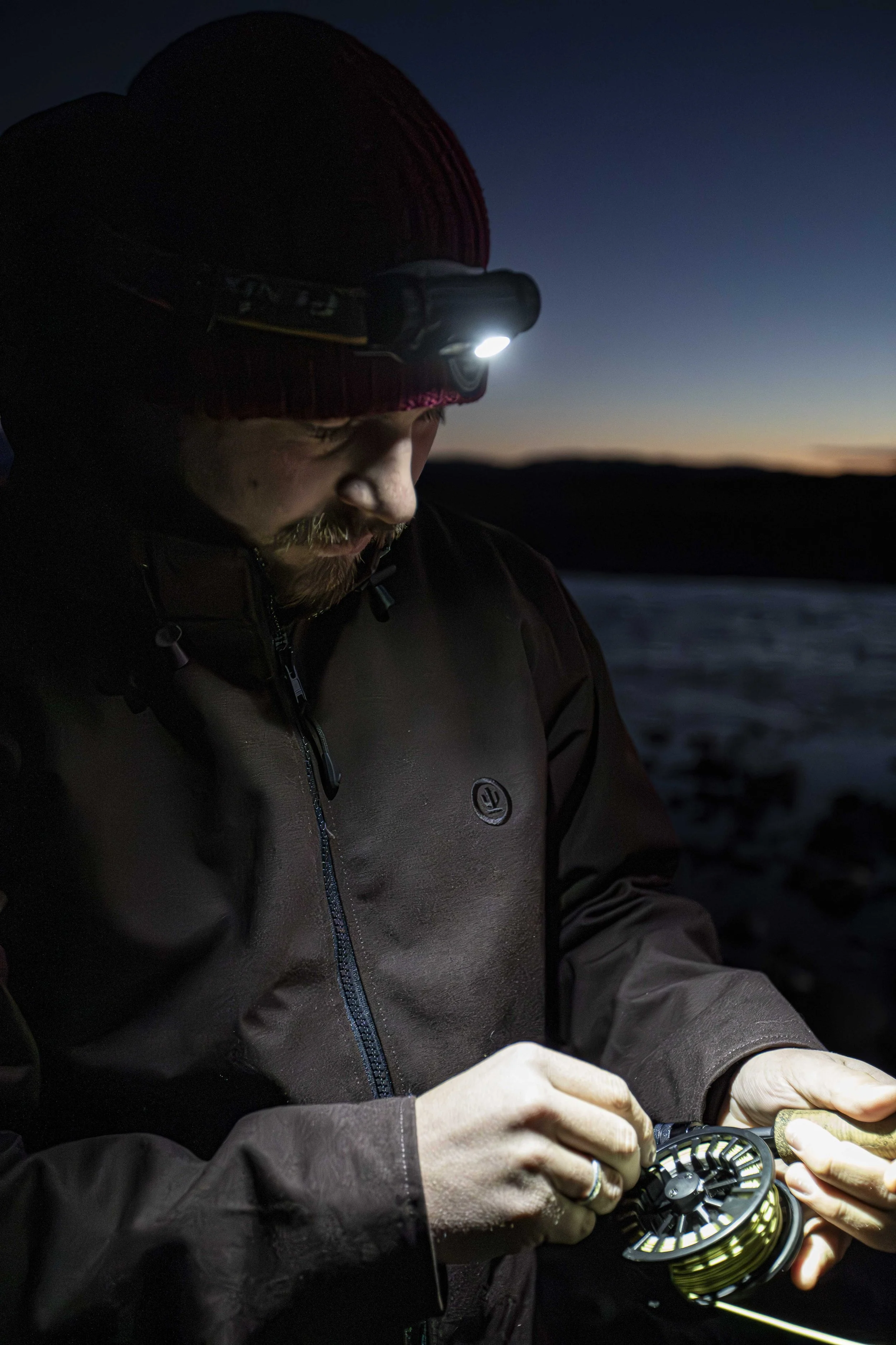 A man wearing a red knit hat, brown jacket, and headlamp, holding a fishing reel outdoors during dusk.