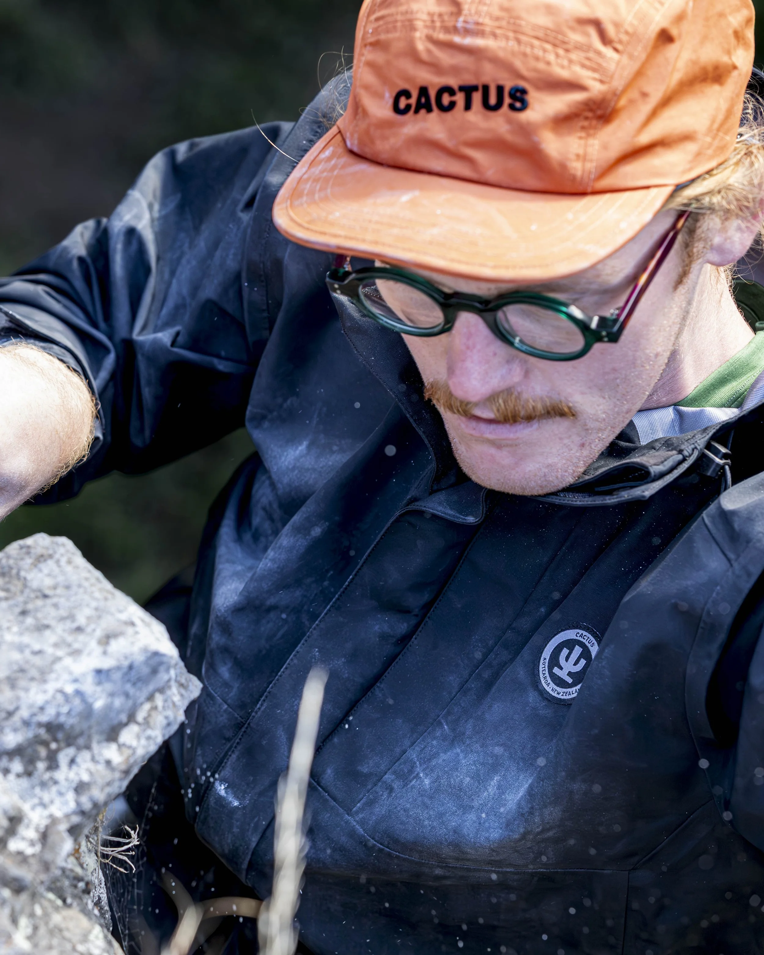 A person wearing an orange cap with the word 'CACTUS', glasses, and outdoor clothing, examining a rock.