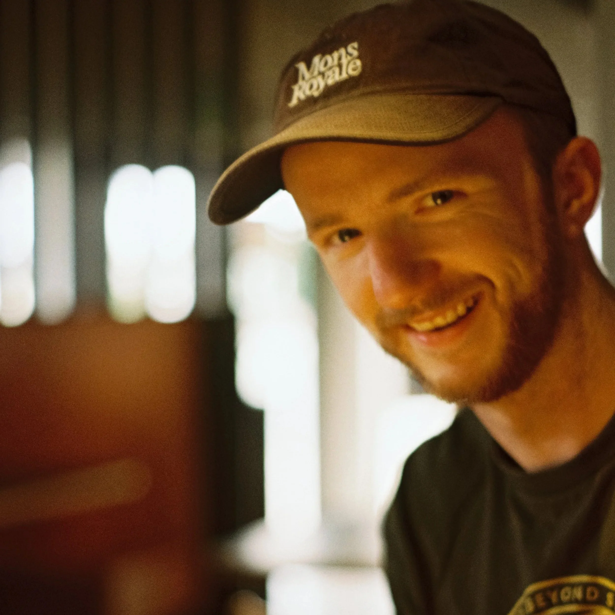 A young man smiling while wearing a brown cap with 'Moms Royale' embroidered on it, and a black shirt.