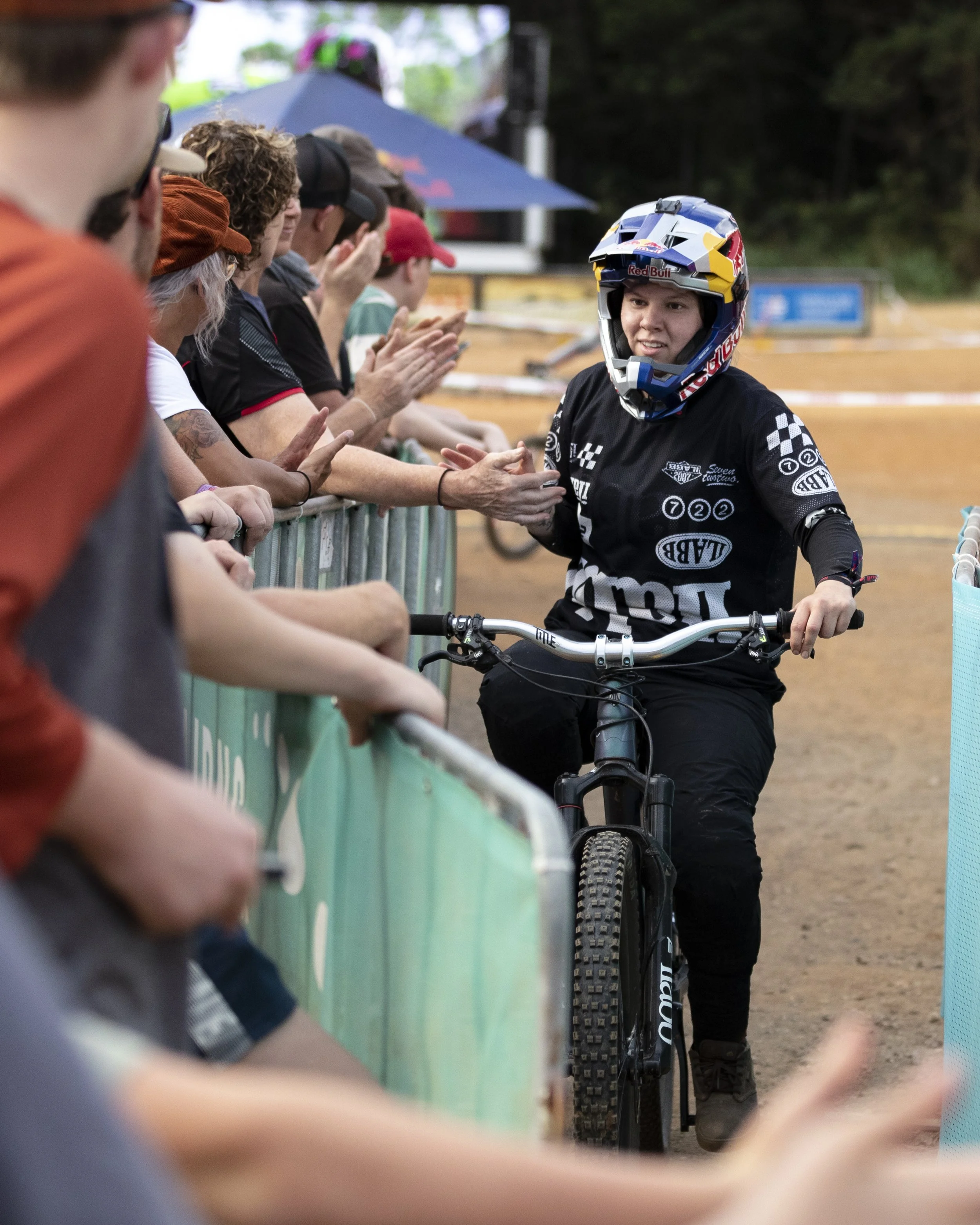 Female mountain biker wearing a helmet and black gear is giving a high five to spectators behind a barricade at a dirt bike trail event.