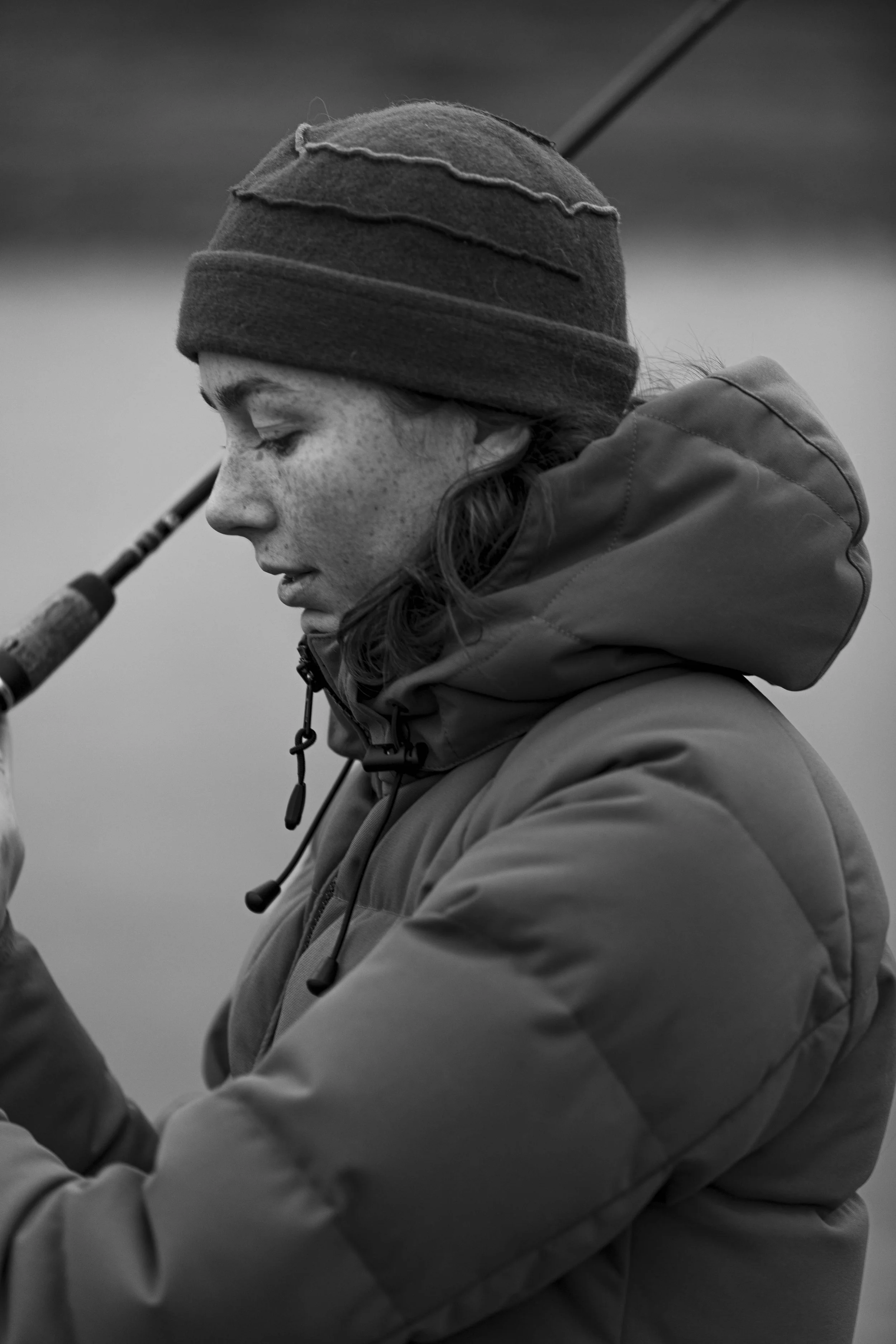 A young woman in a winter jacket and beanie hat looks down as she holds fishing gear. Black and white photo.