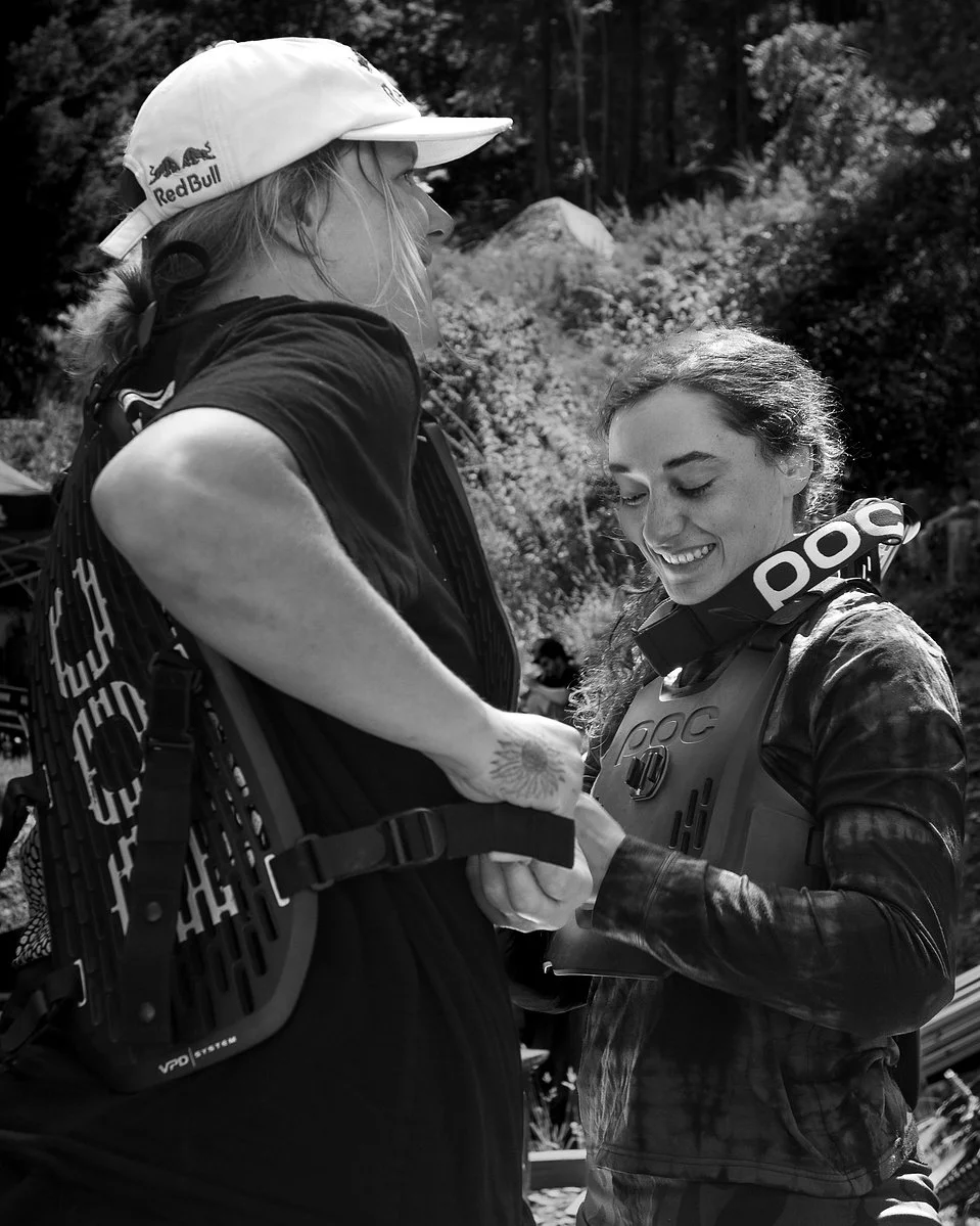 Two women outdoors, one is helping the other by fastening her motorcycle armor. The woman on the left is wearing a White cap with a Red Bull logo and a black t-shirt; the woman on the right is wearing a black jacket and protective gear with the brand name 'POC' visible.