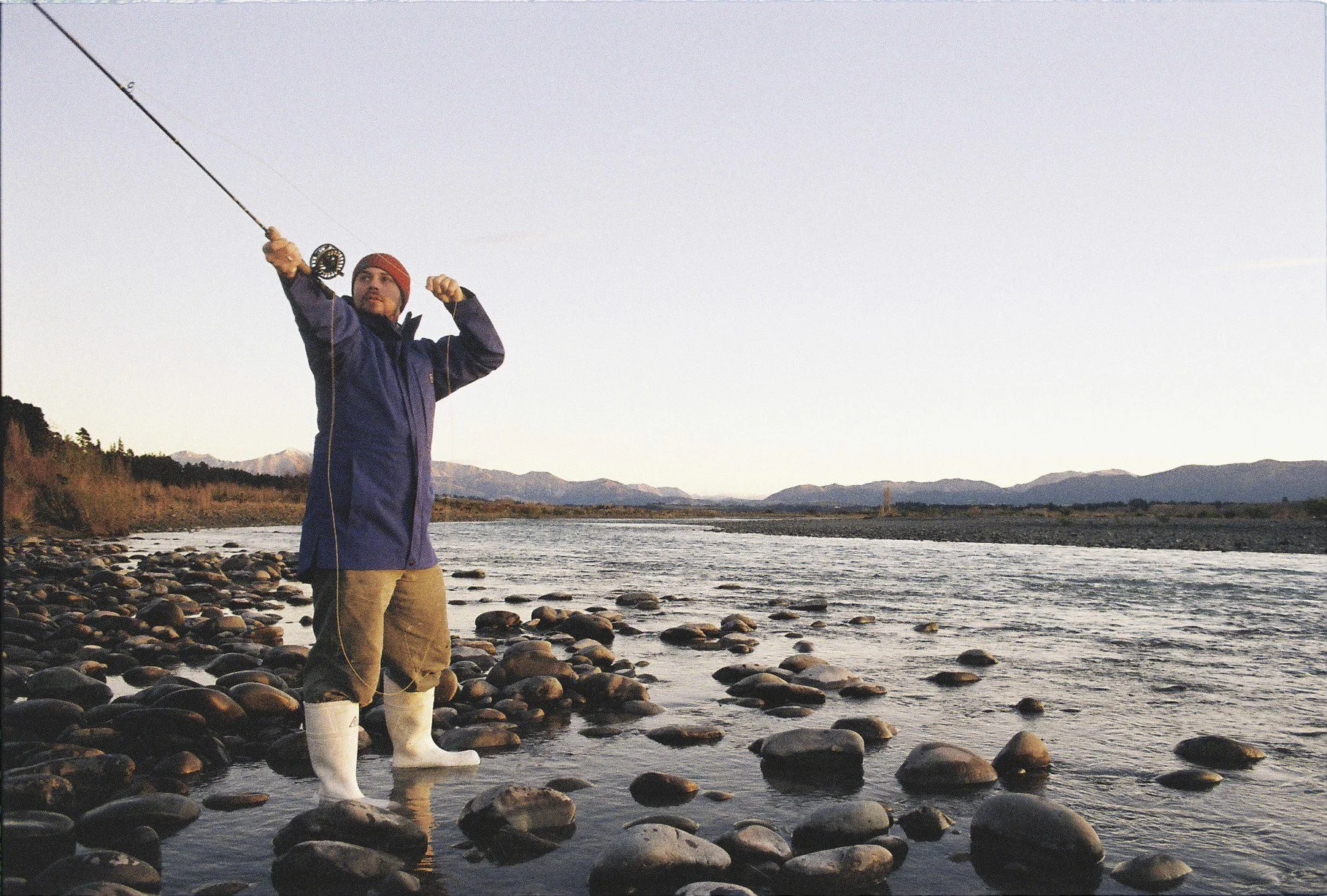 A man standing in a rocky riverbank casting a fishing line during sunset.