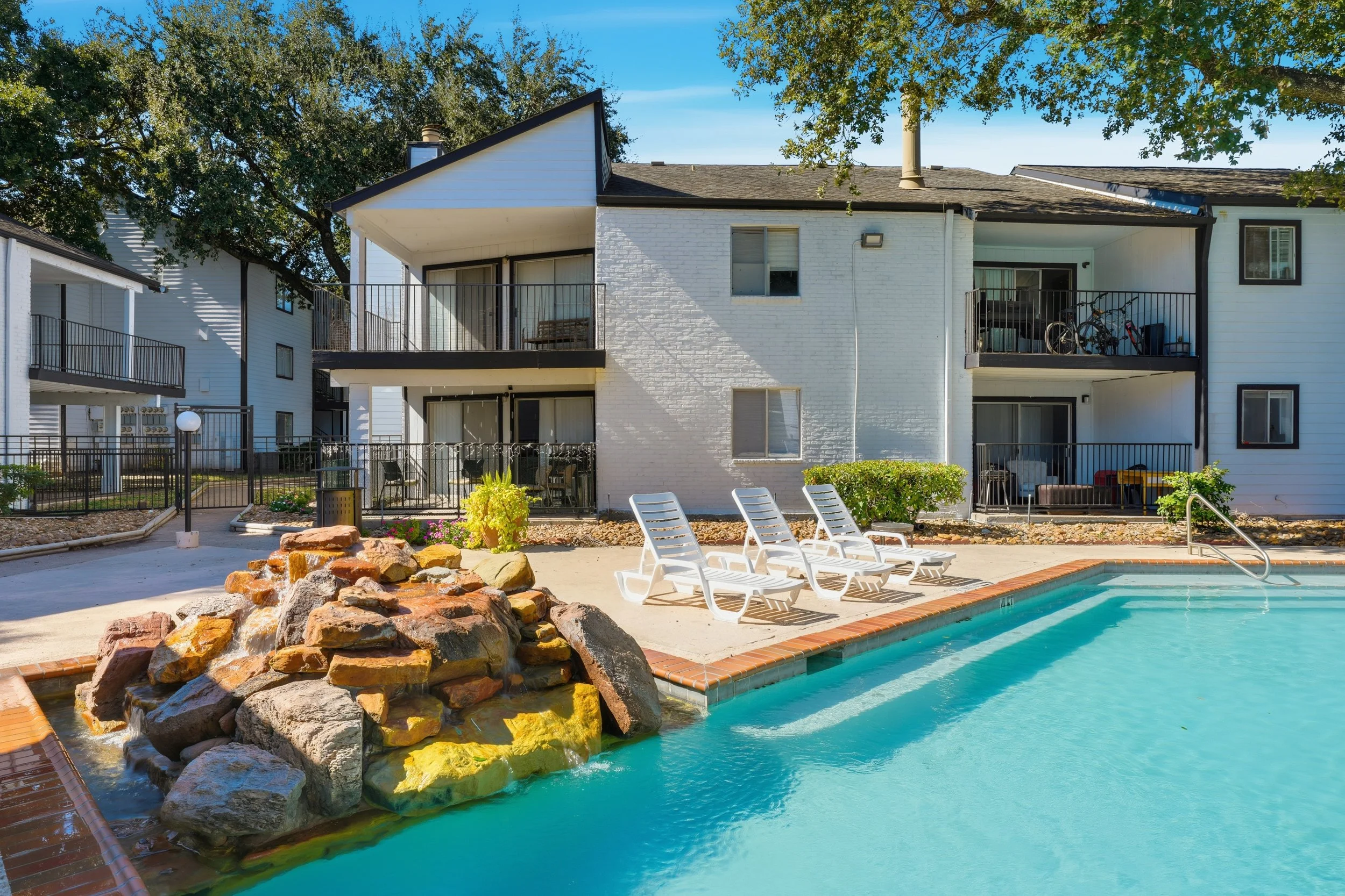 Outdoor swimming pool with white lounge chairs and a rock waterfall feature, apartment building with balconies, and surrounding trees