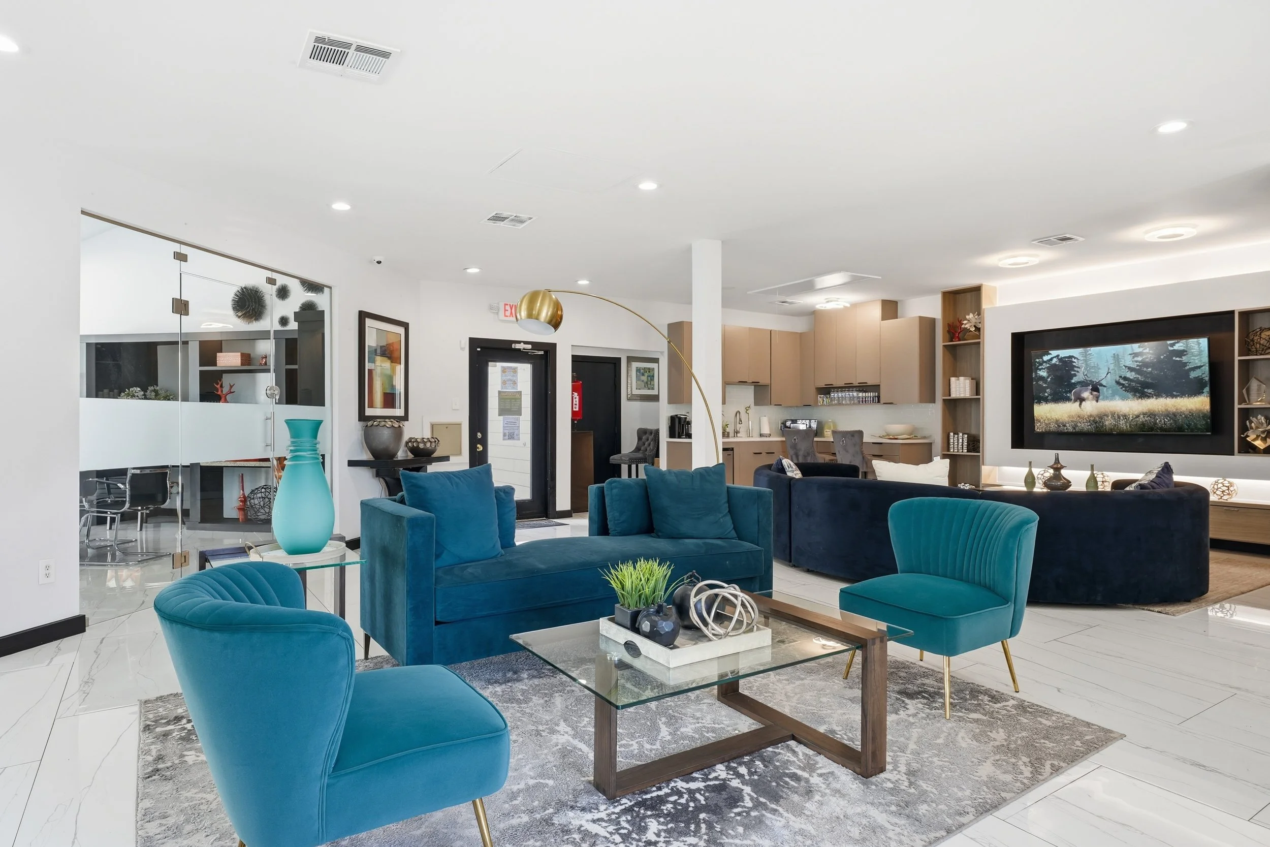 Modern lobby or lounge area with blue velvet chairs and sofa, a glass coffee table with decorative items, a large wall-mounted TV displaying nature scene, and a kitchen area in the background.