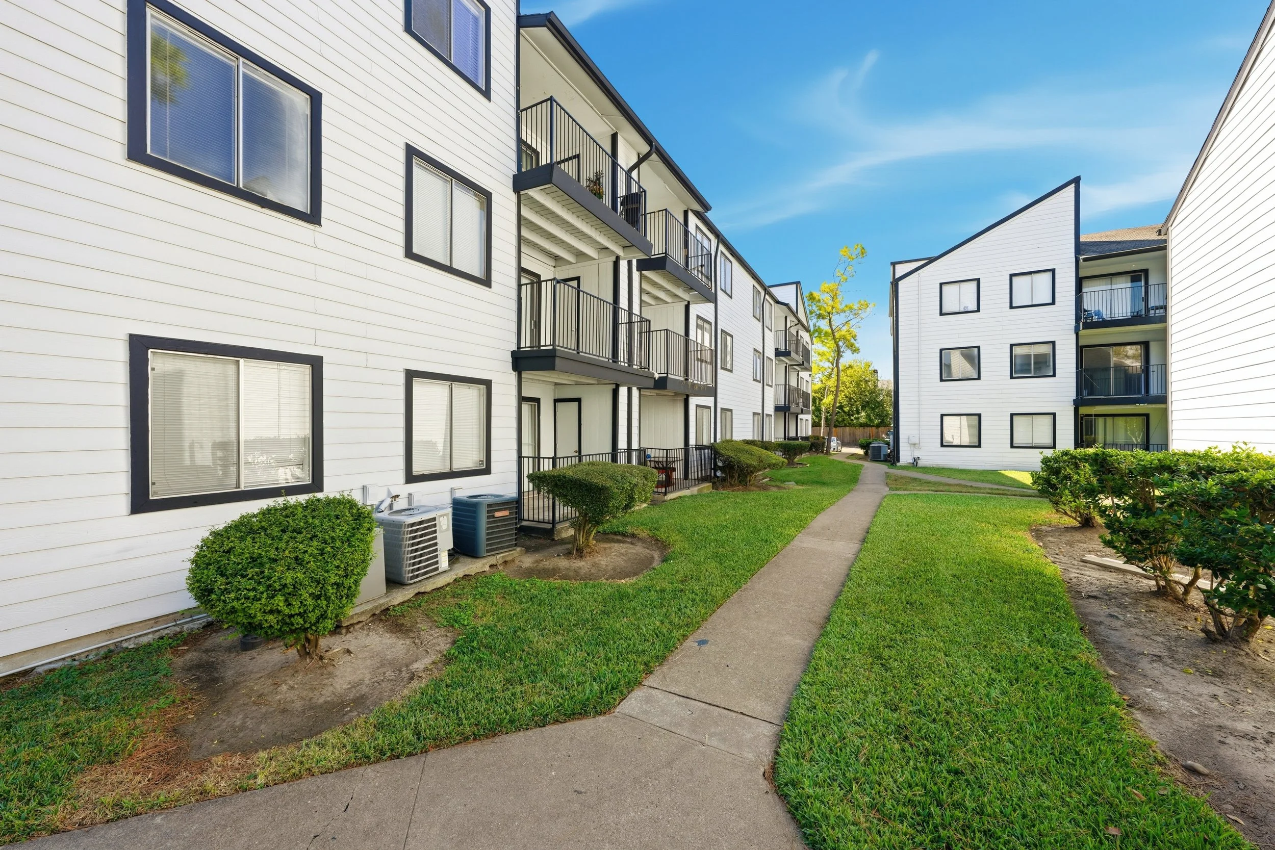 Exterior view of a white apartment complex with multiple buildings, black railings on balconies, green lawn, paved walkway, and some shrubs and bushes under a clear blue sky.