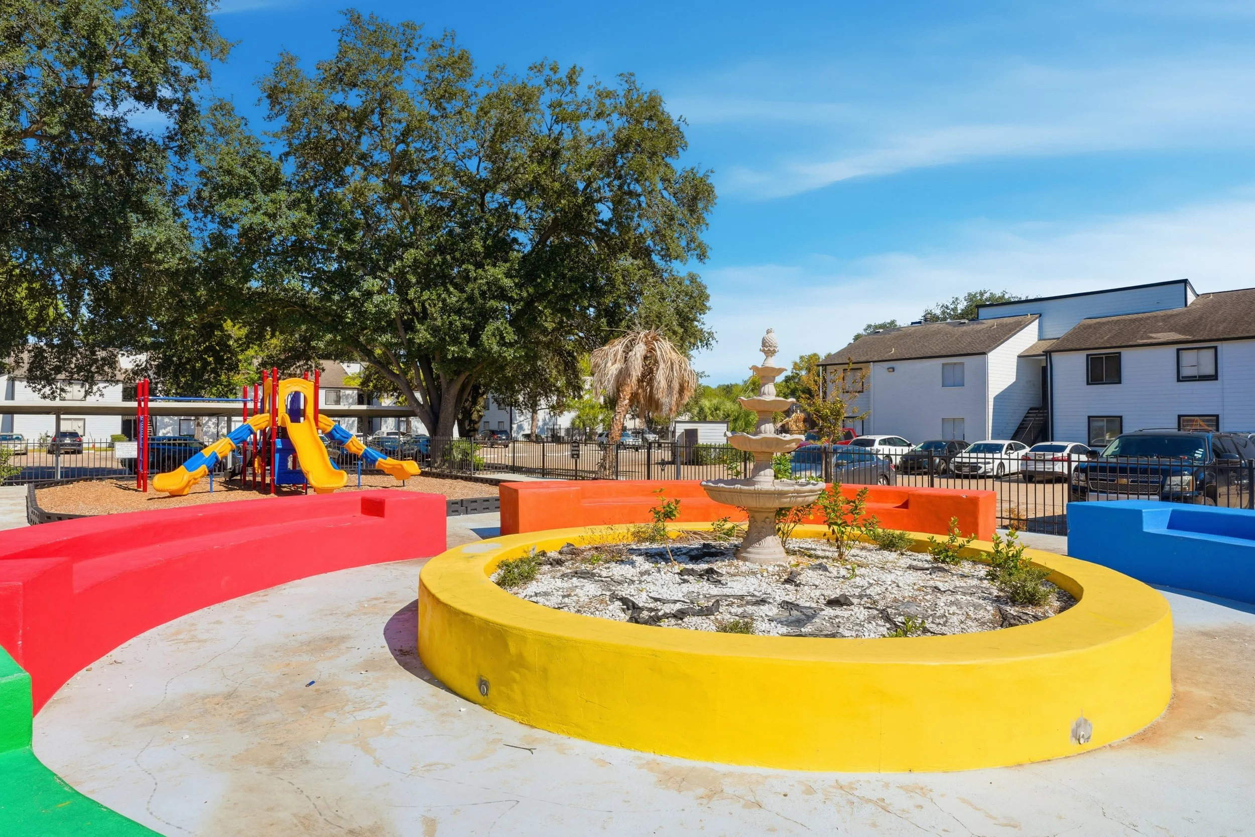 Colorful playground area with slides and climbing structure, surrounding a decorative fountain on a sunny day.