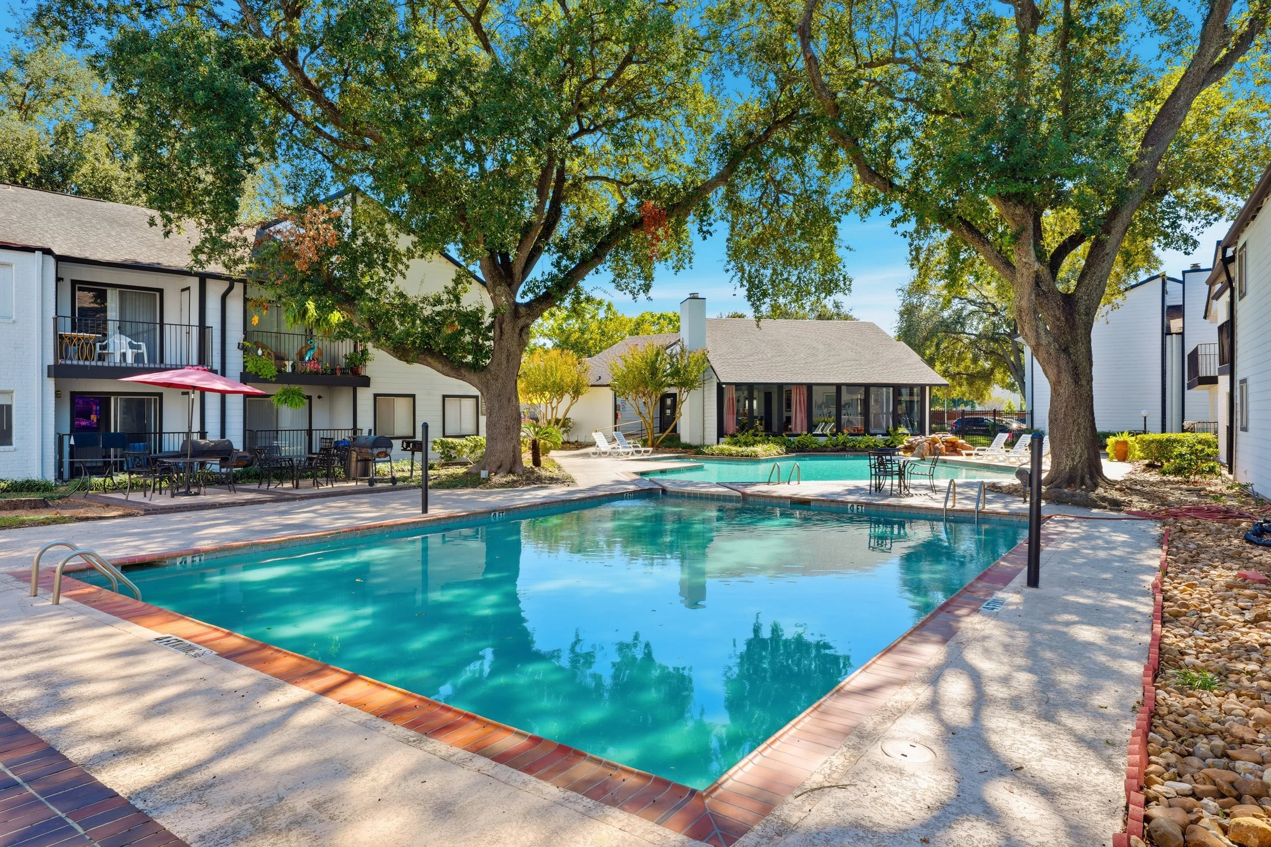 Two swimming pools with lounge chairs, surrounded by trees and apartment buildings on a sunny day.