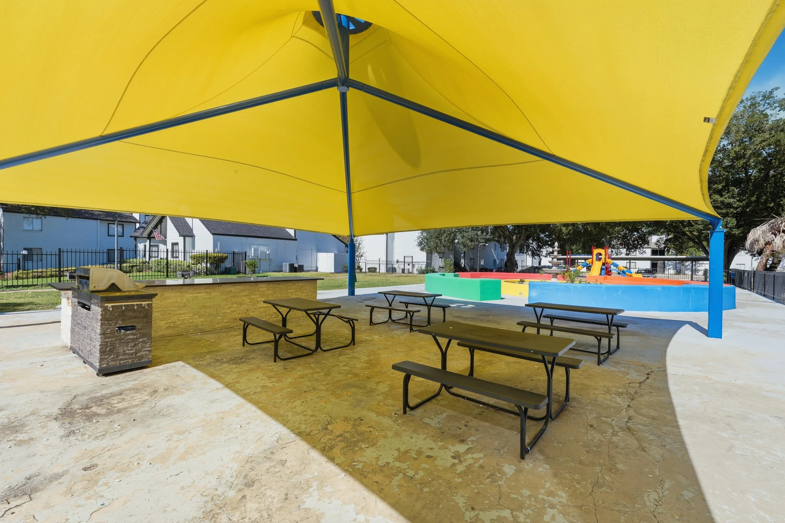 Children's playground with a yellow canopy, picnic tables, a sandbox with colorful walls, and play structures, all under a sunny sky.