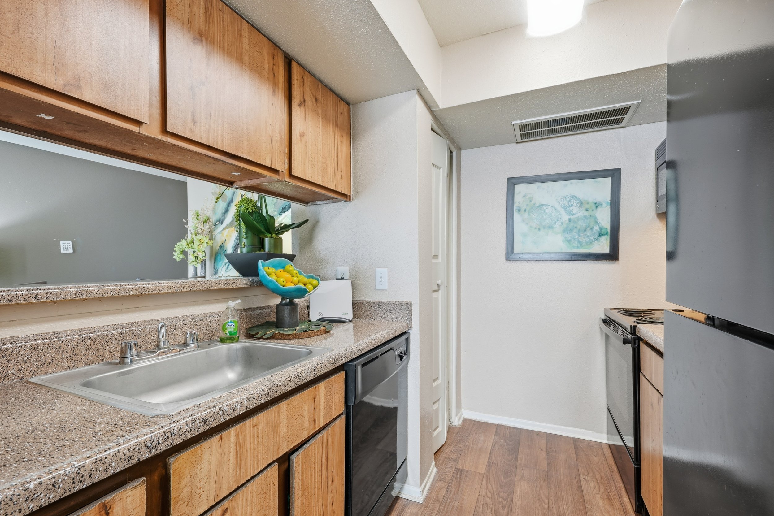 Kitchen with wooden cabinets, beige speckled countertop, stainless steel sink, black stove, and a refrigerator. Decor includes plants, a bowl of lemons, and a framed picture on the wall.