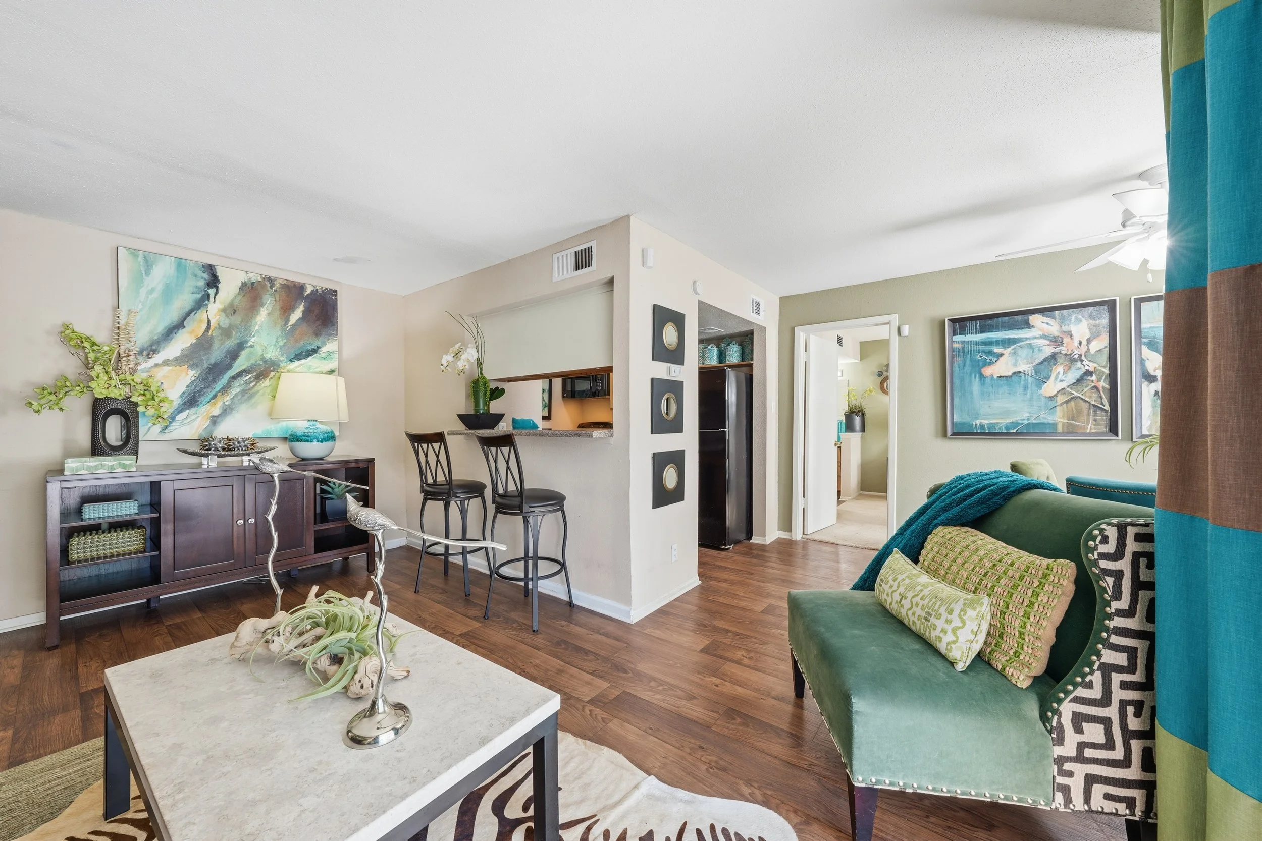 Living room with green sofa, colorful pillows, artwork, wooden floor, and a partial view of an adjacent kitchen with a breakfast bar and barstools.