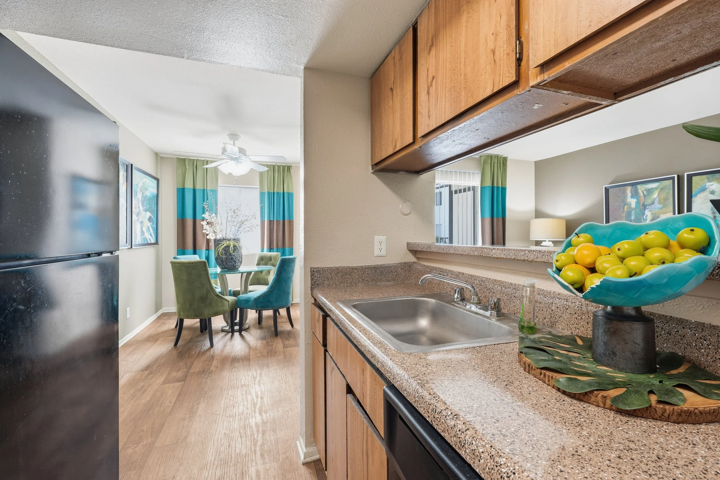 Kitchen with a beige speckled countertop, a bowl of green apples on a black pedestal, and an under-mount stainless steel sink. In the background, there's a dining area with a round table surrounded by four colorful chairs, behind a window with green,