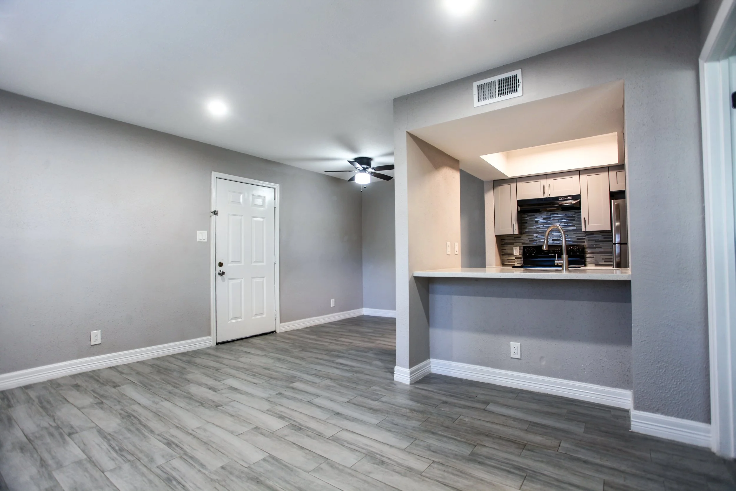Empty living room with gray walls, white door, wood-like laminate flooring, and visible kitchen area with gray cabinets and a tile backsplash.