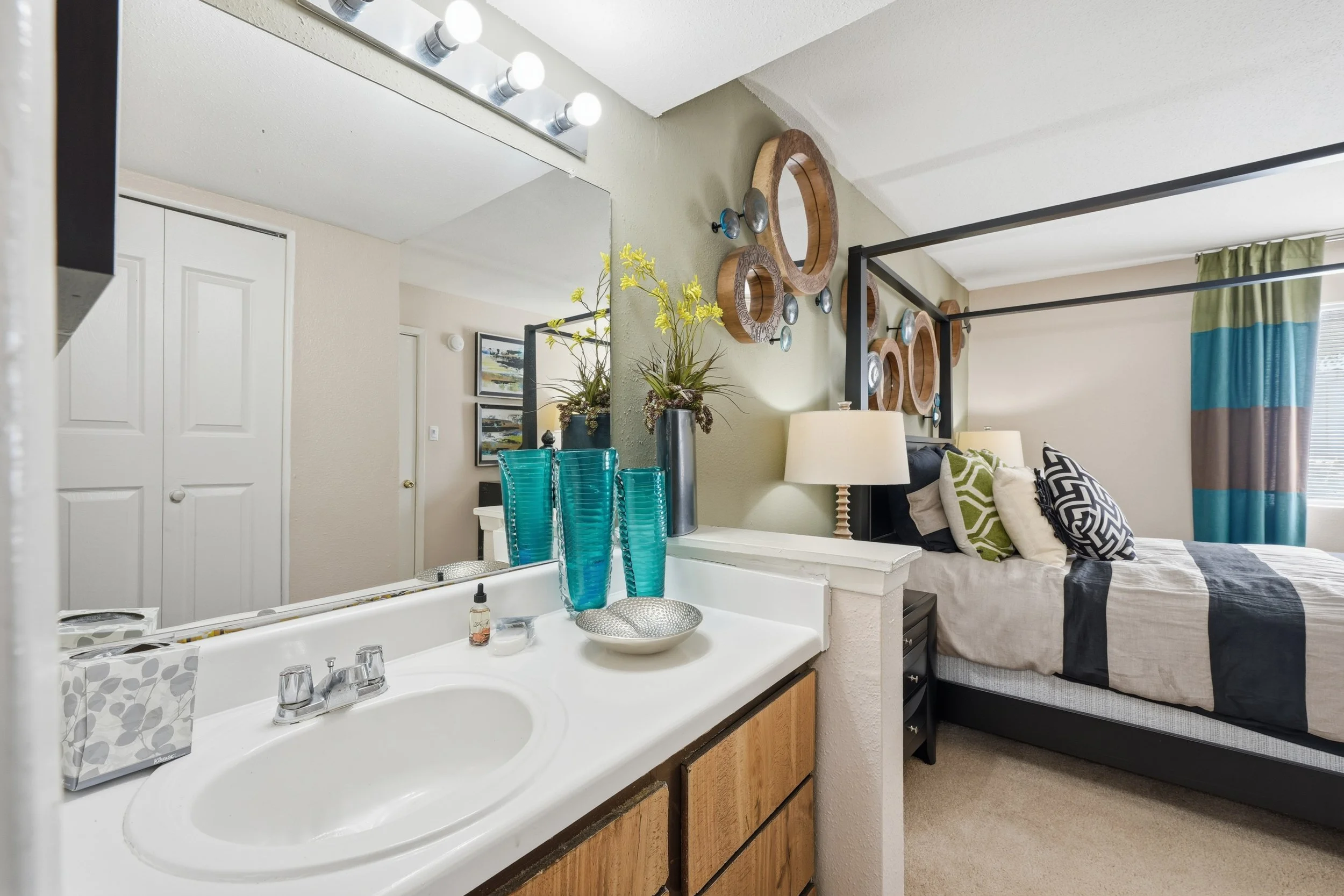 A bedroom with a bed and colorful pillows, a nightstand with a lamp, wall decorations, and a window with curtains. Adjacent to the bedroom is a bathroom sink area with turquoise vases, a mirror, and decorative items.