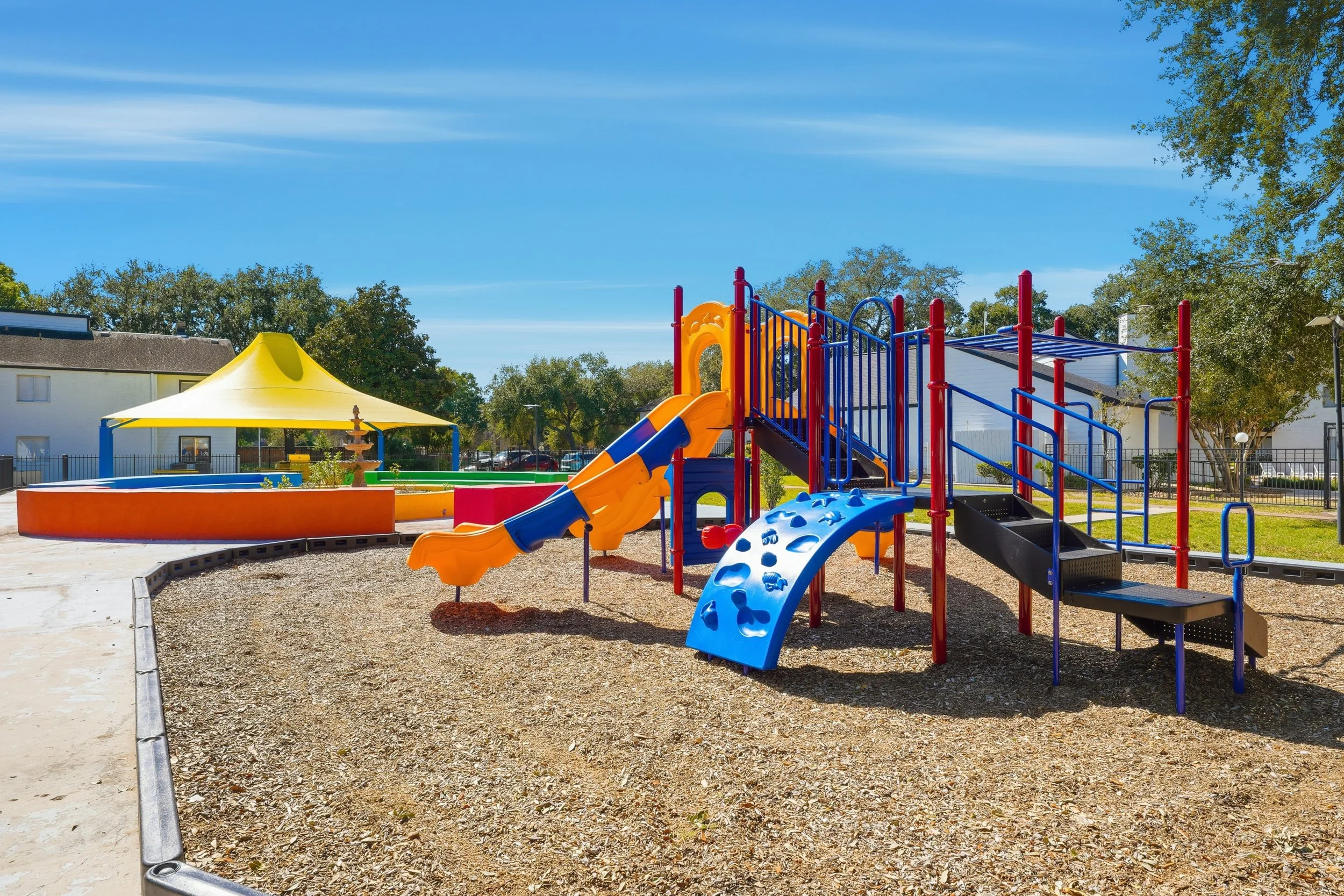 Colorful playground with slides, climbing structures, and a shaded area on a sunny day with trees and houses in the background.