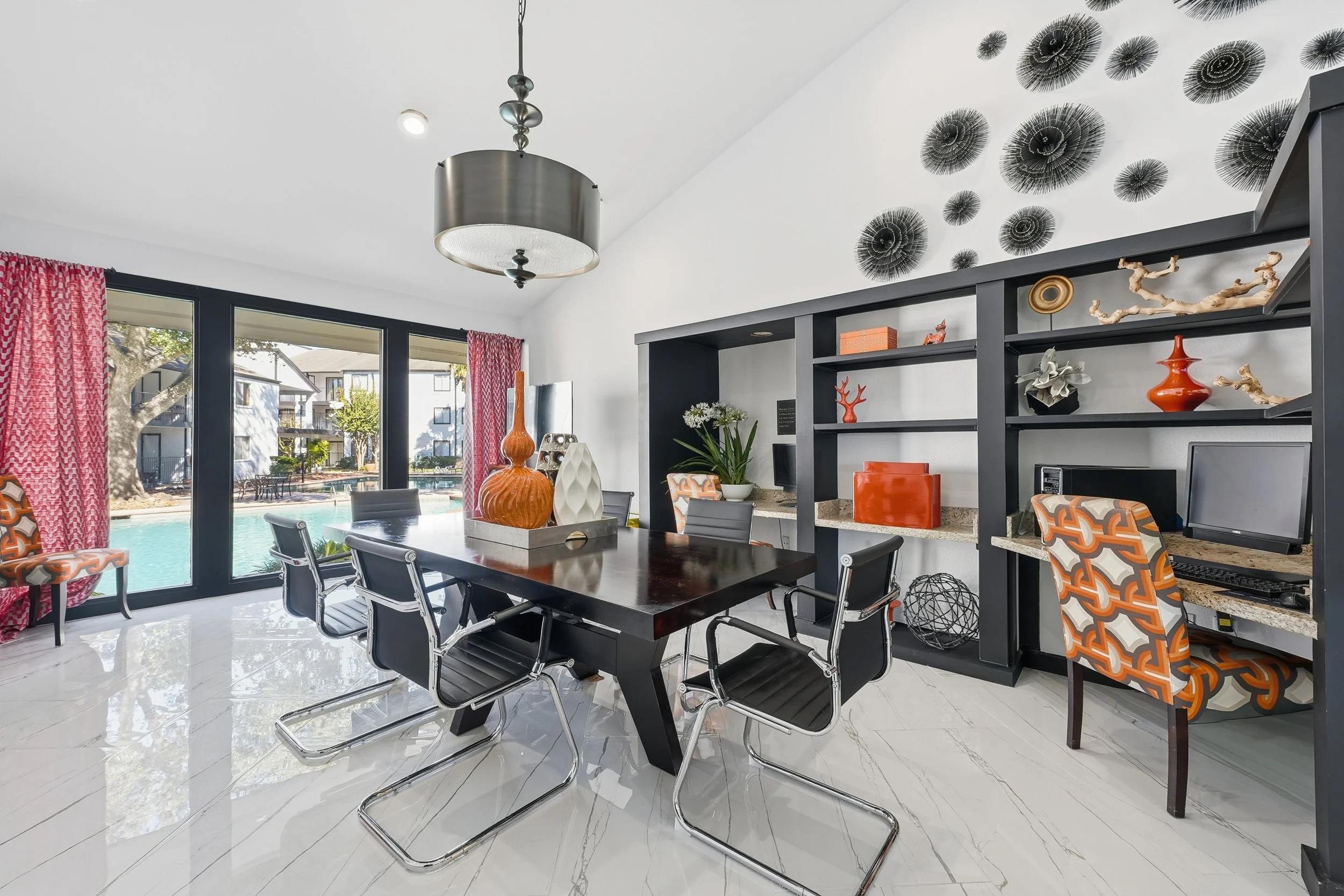A modern dining room with a black dining table and six black chairs with chrome frames. Large windows with red patterned curtains overlook a pool and outdoor area. A black built-in shelf displays decorative items, and a black pendant light hangs from