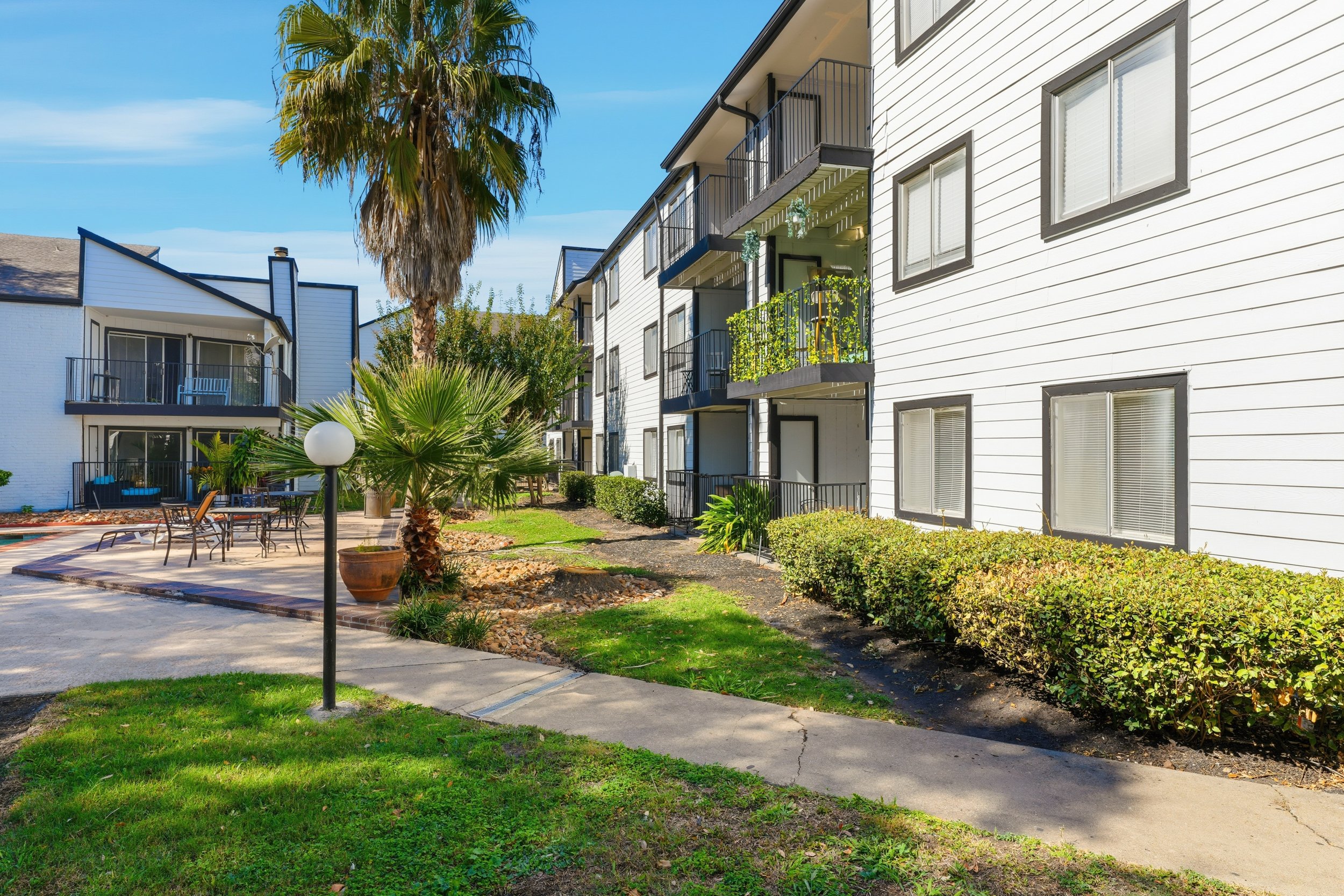 Exterior view of a white apartment building with balconies, surrounded by palm trees, bushes, and a lawn, on a sunny day.