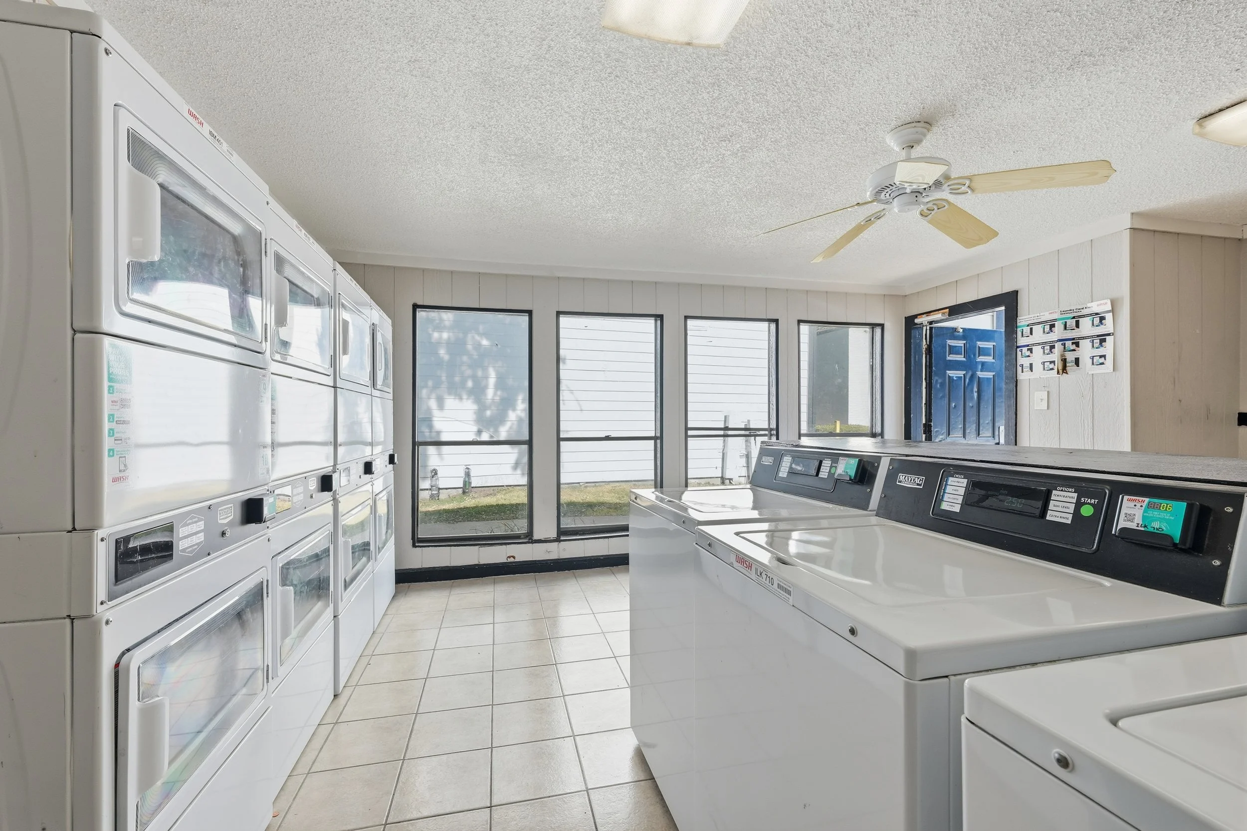 Laundry room with stacked washers and dryers, tiled floor, sliding glass windows, ceiling fan, and blue door.