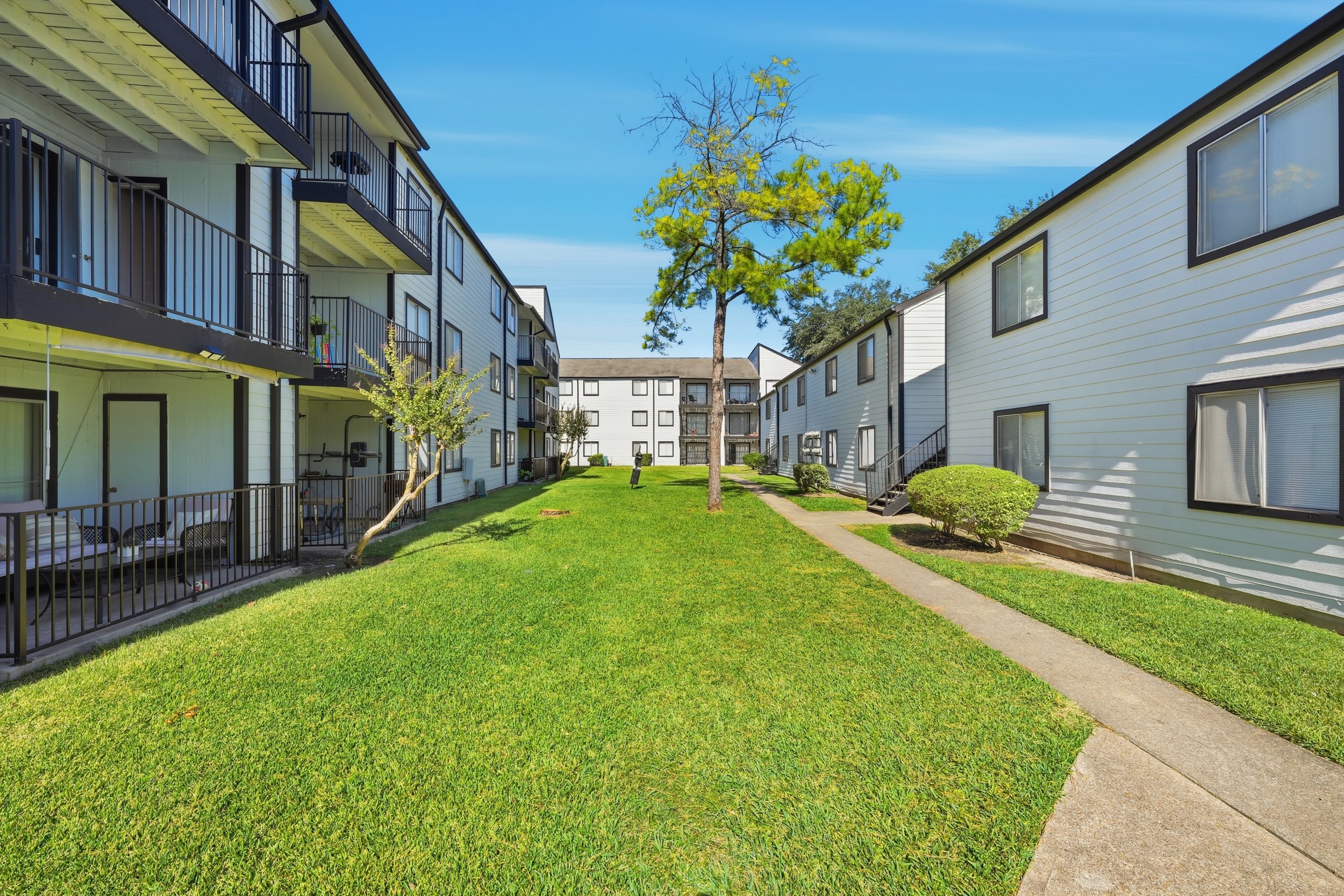 Apartment complex with white buildings, green lawn, paved walkways, and several trees under a blue sky.