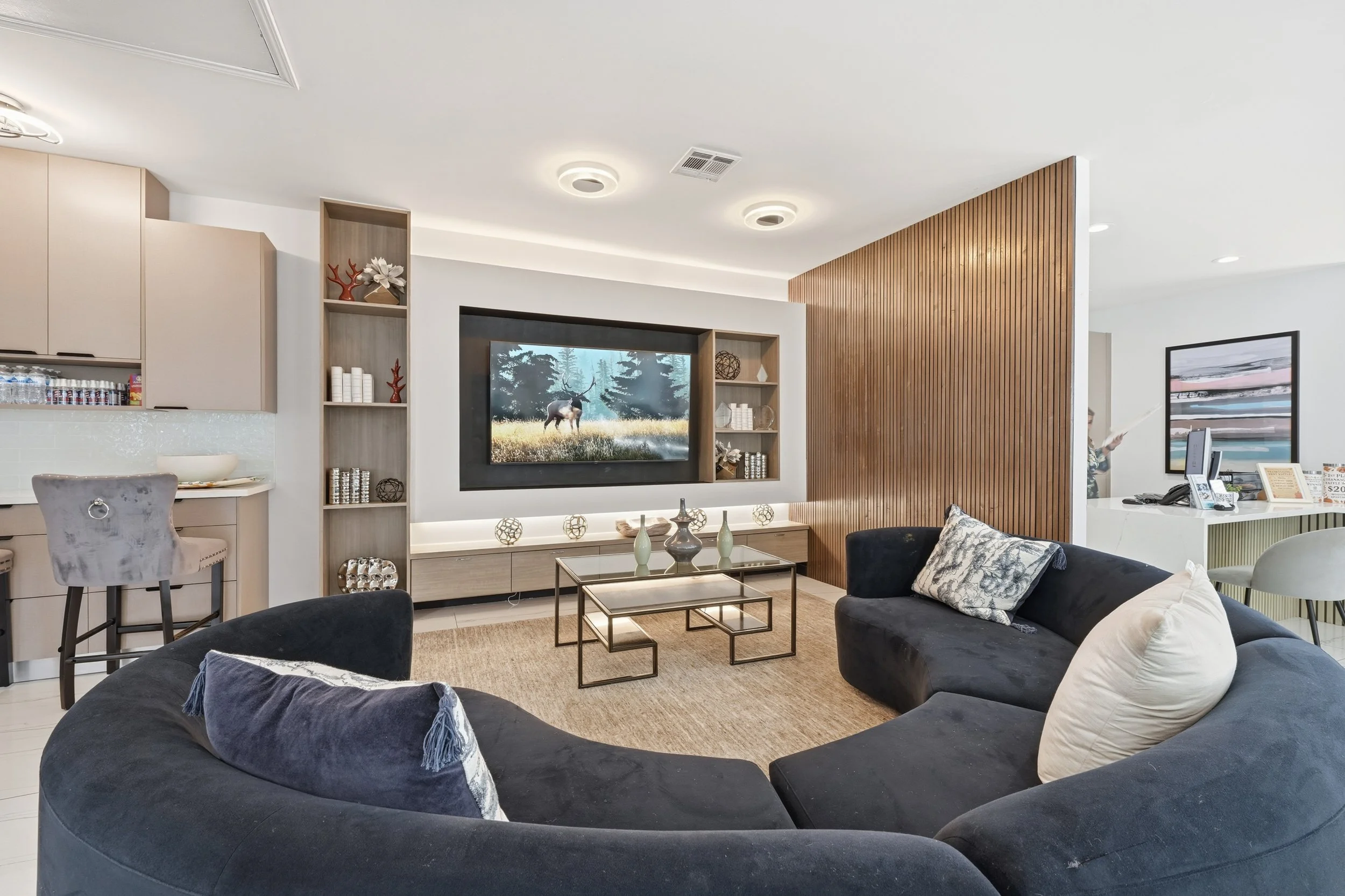 A modern living room with black curved sofas, decorative cushions, a glass coffee table with vases, a television on a wall shelf, beige kitchen cabinetry, and a person at a reception desk in the background.
