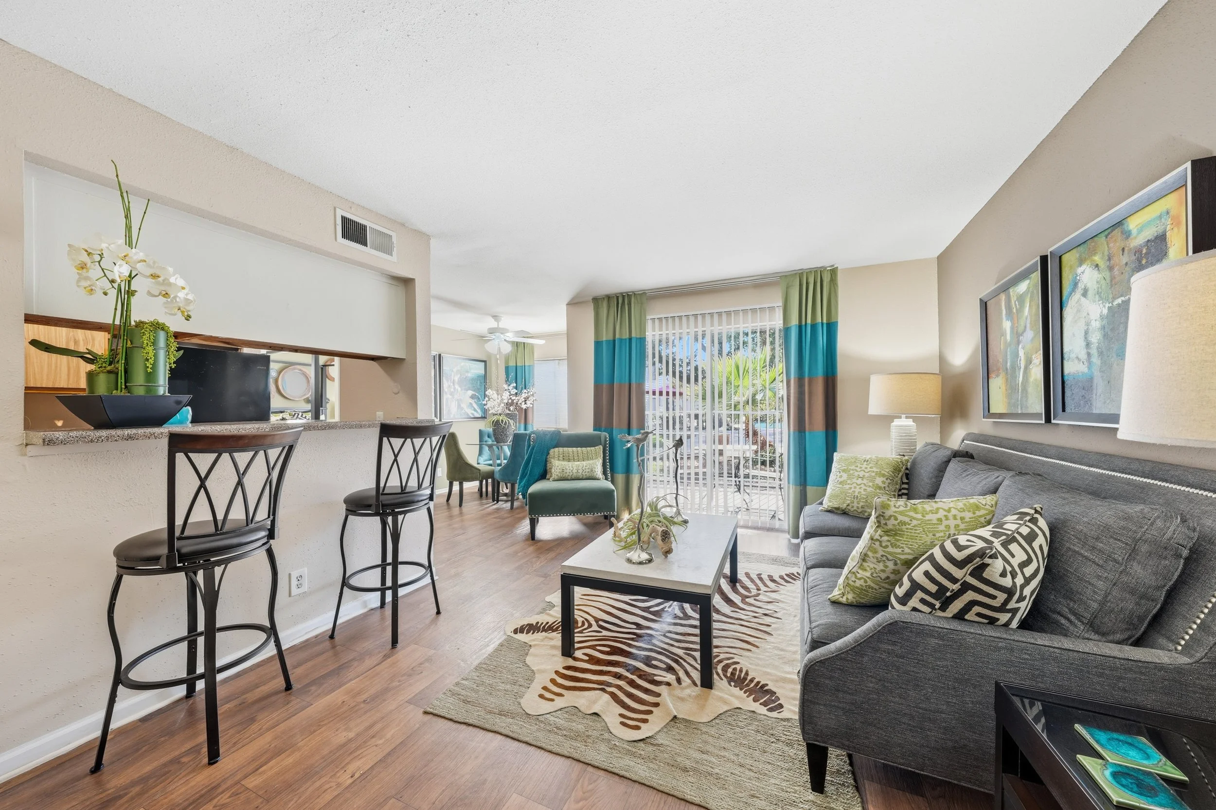 Living room with gray couch, green and beige pillows, two table lamps, artwork on the wall, wooden flooring, curtains with green, blue, and brown stripes, sliding glass door leading to patio, and a dining area with chairs and a table near the window.