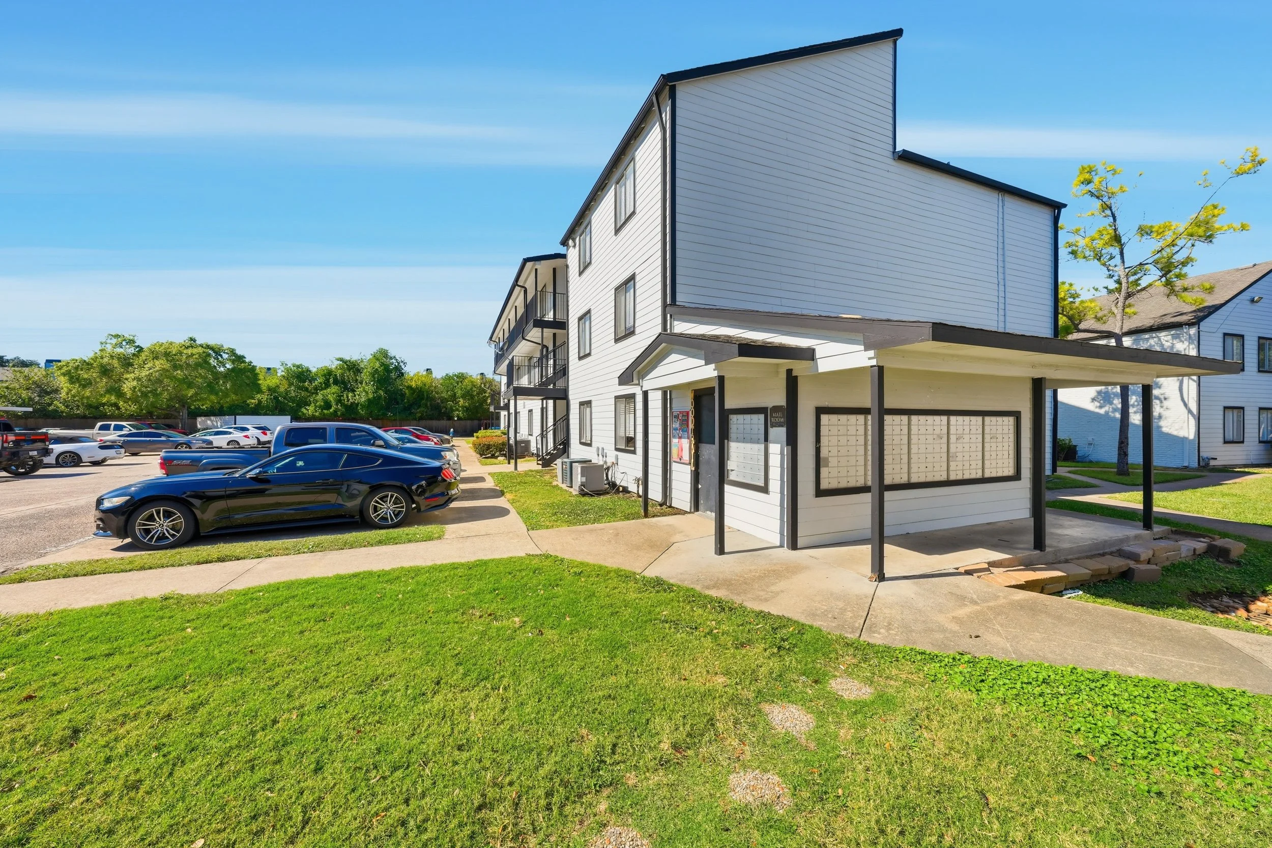 A multi-story white apartment building with a small covered parking area and adjacent parking lot filled with cars, with a grassy lawn in the foreground and trees in the background under a blue sky.
