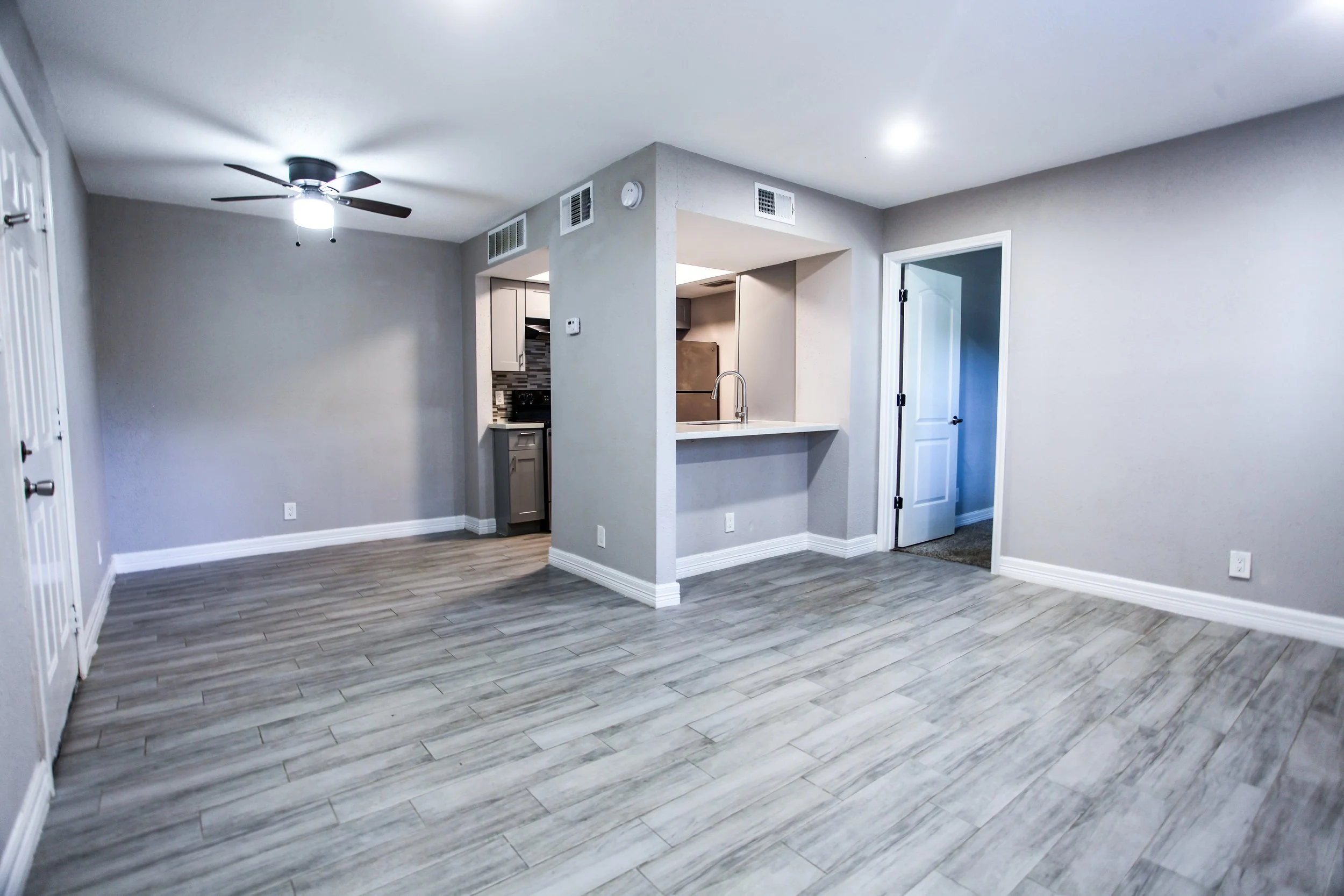 Empty living room with light gray walls and wood-like floor tiles, ceiling fan, and open kitchen with granite countertops and gray cabinets.