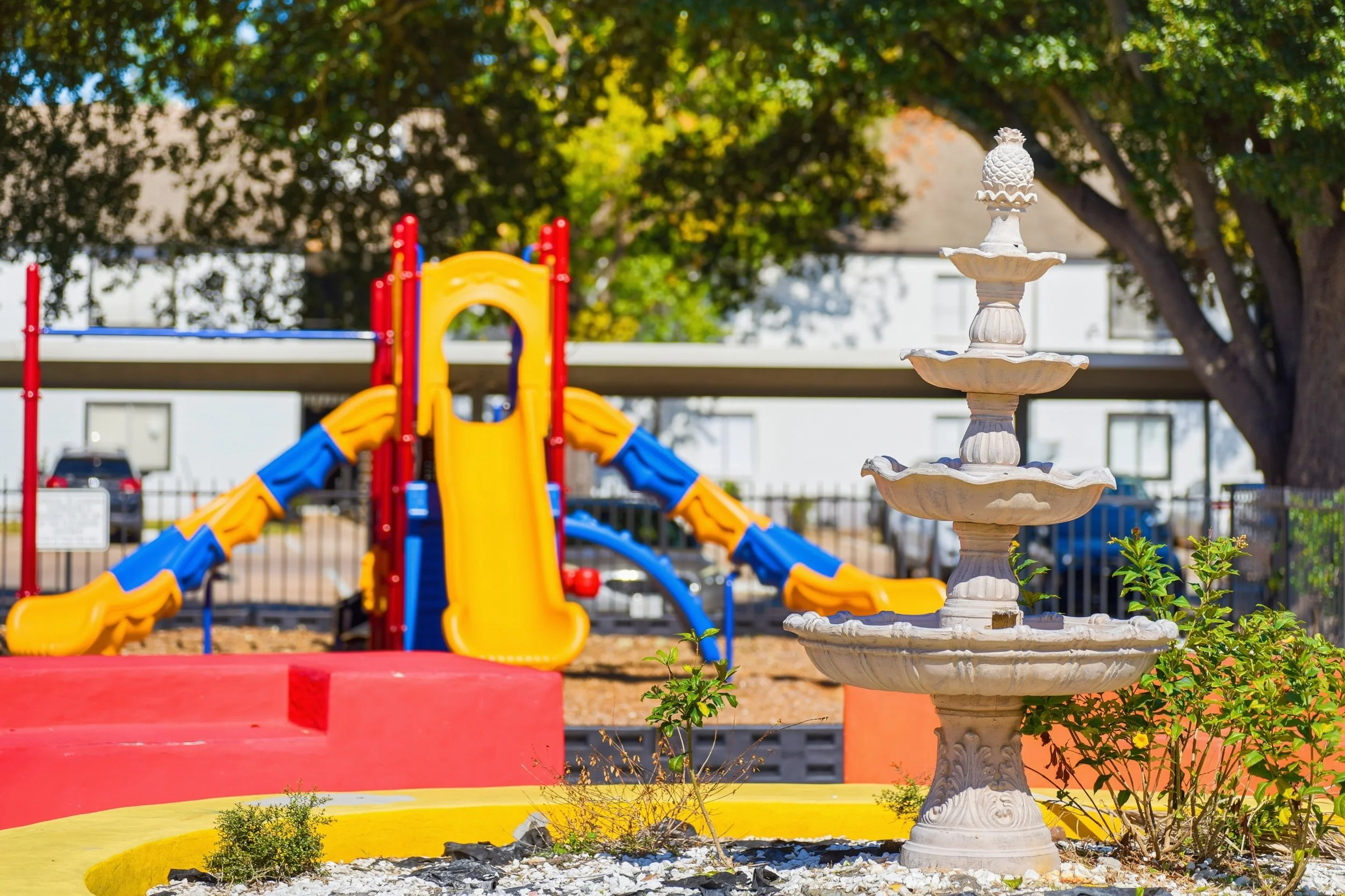 Colorful playground with yellow, red, and blue slides and a three-tier white fountain in the foreground, surrounded by small green plants and trees in the background.