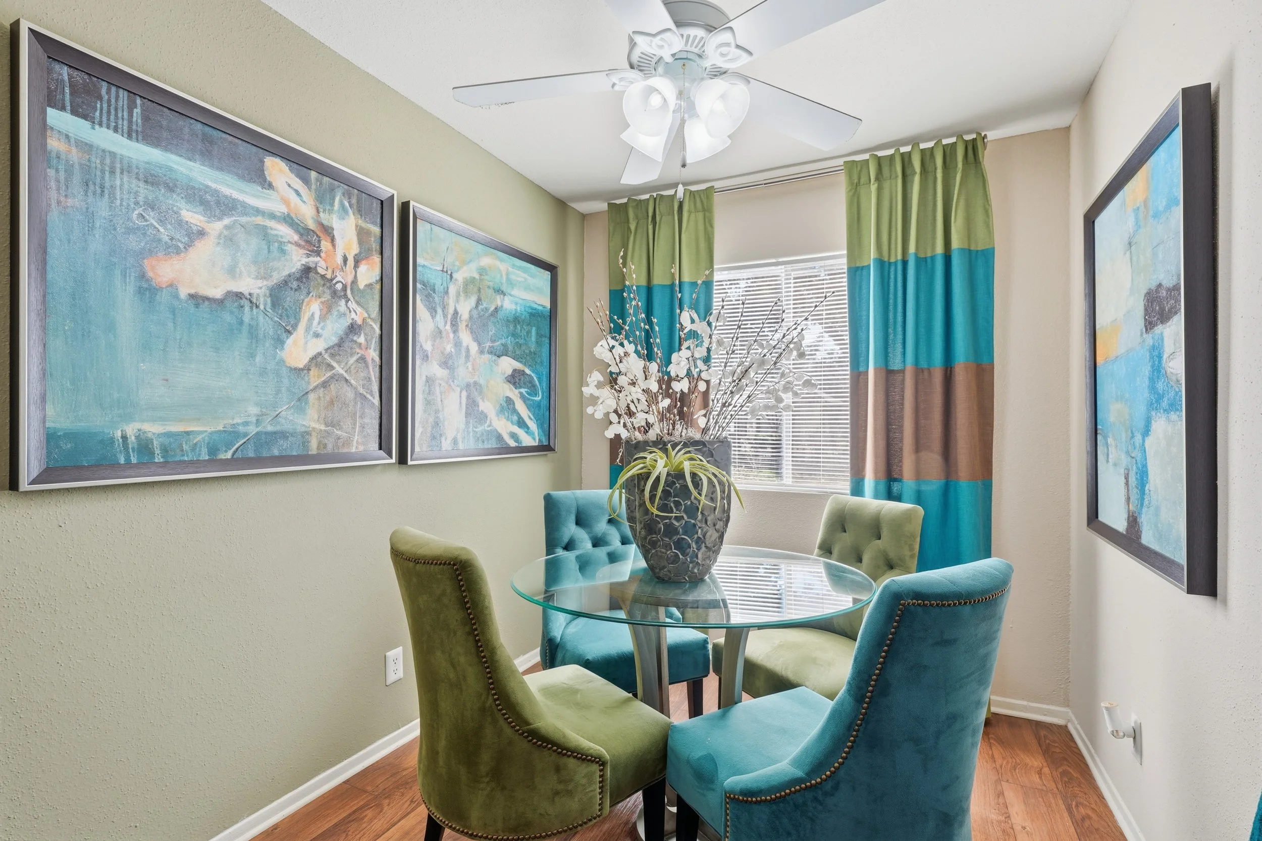 A dining room with a round glass table surrounded by six upholstered chairs in shades of blue and green. A large decorative vase with branches and white flowers is on the table. The room has light green and beige walls, framed abstract artwork, green
